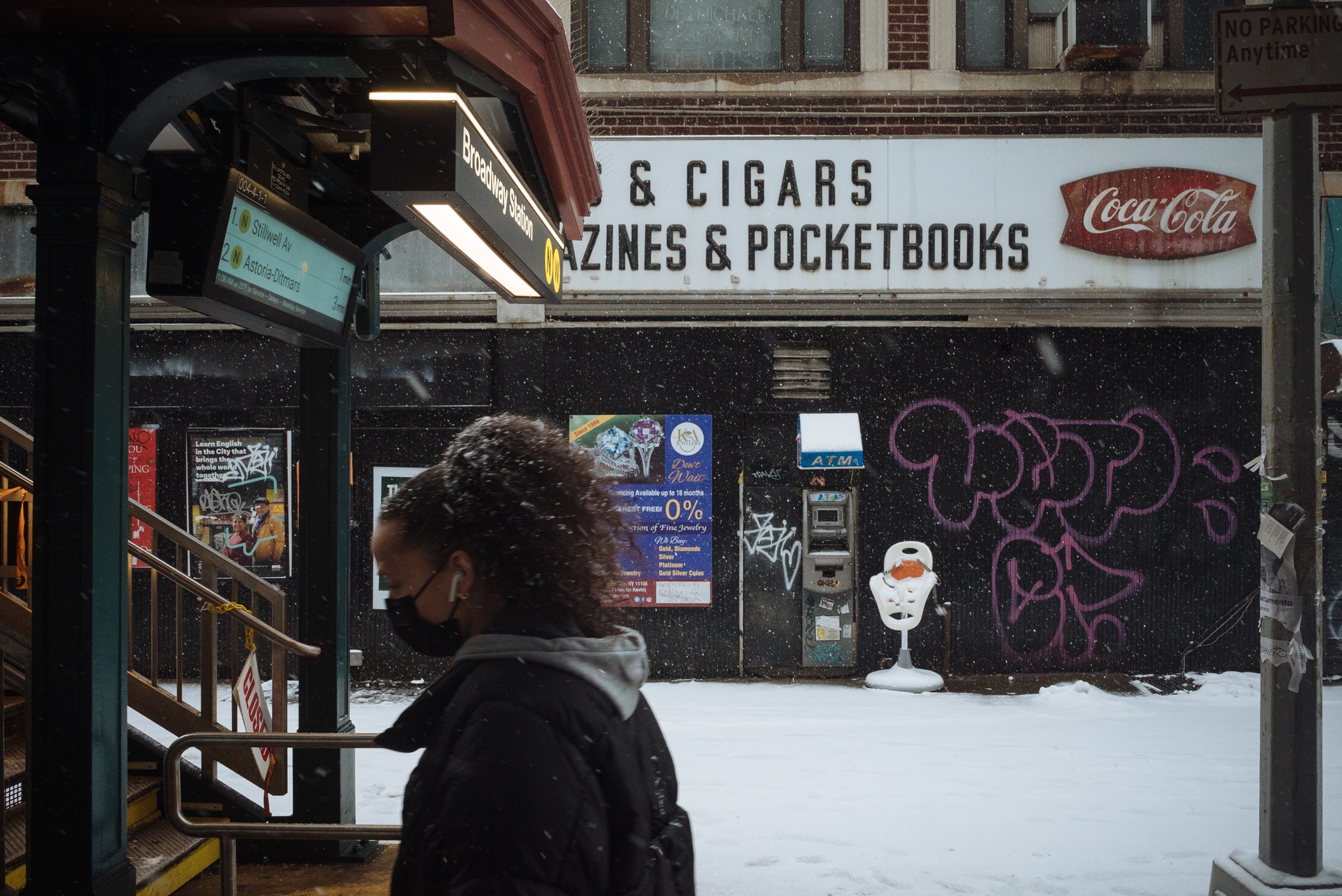 A person in a mask walks past a subway entrance in a snowy city. Behind them, store signs advertise cigars, magazines, and pocketbooks. Graffiti and an ATM are visible on the wall.