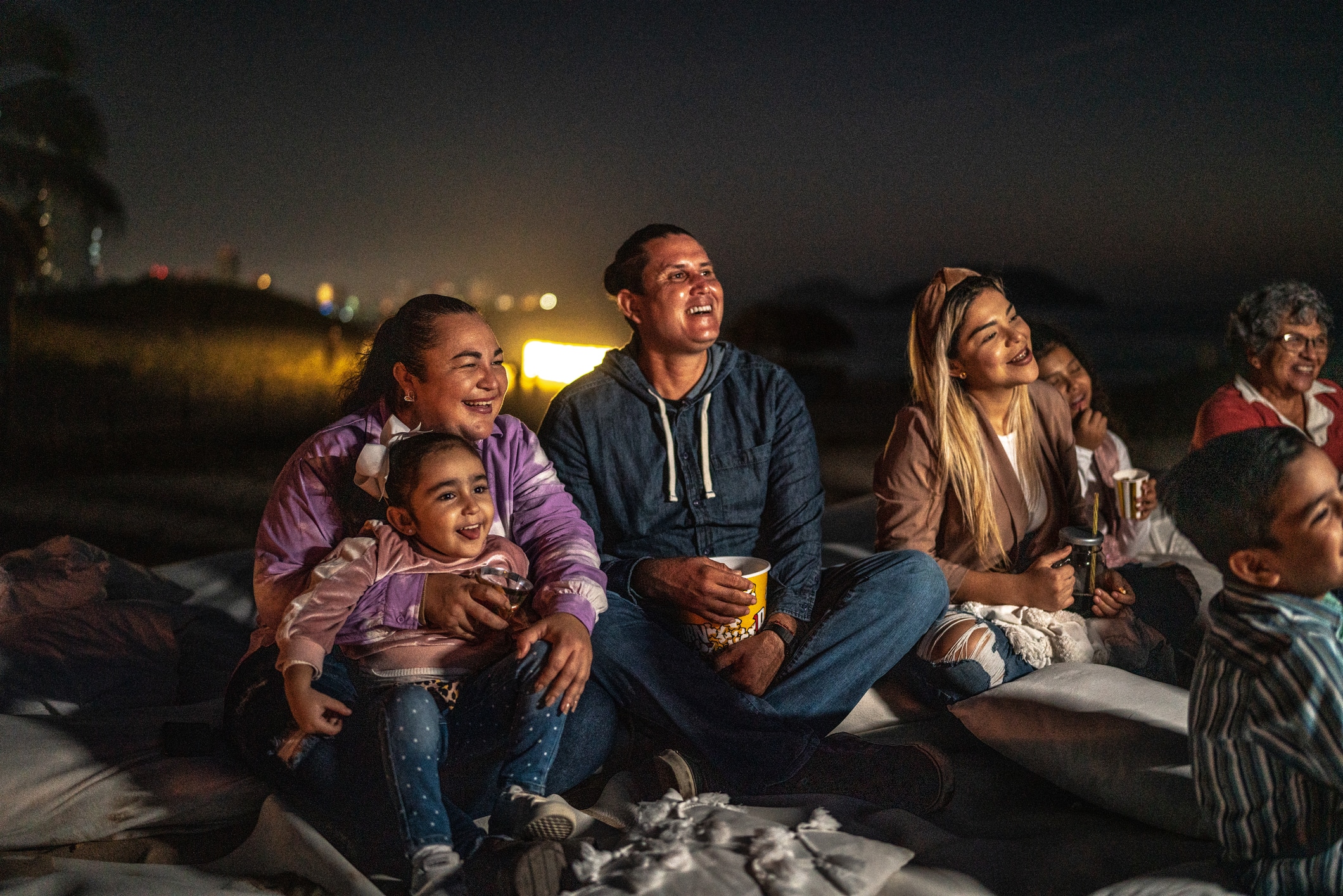A group of people, including children and adults, sit together on cushions outdoors at night, smiling and watching something off-camera. Some are holding popcorn and drinks. The background is dark with blurred lights.