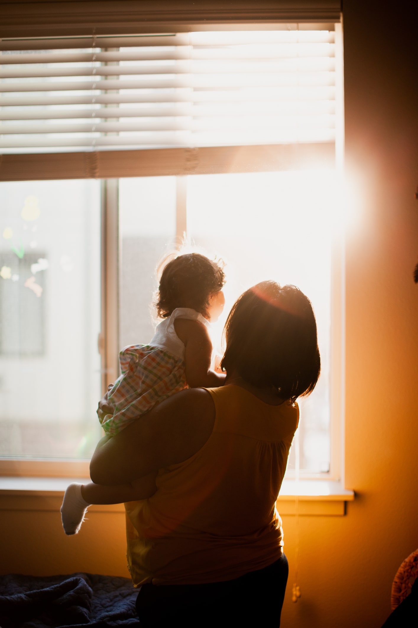 An adult holds a child while standing in front of a sunlit window with partially open blinds. Both figures are in shadow, facing the bright sunlight coming through the window.