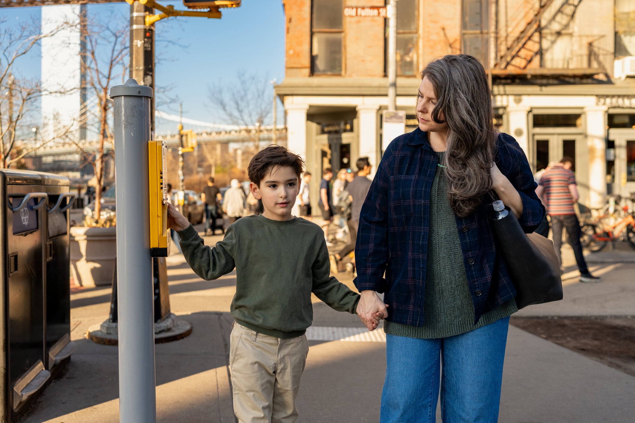 A woman and a young boy holding hands wait at a crosswalk. The boy is pressing the button. They are on a city sidewalk, with people, buildings, and a bridge visible in the background.