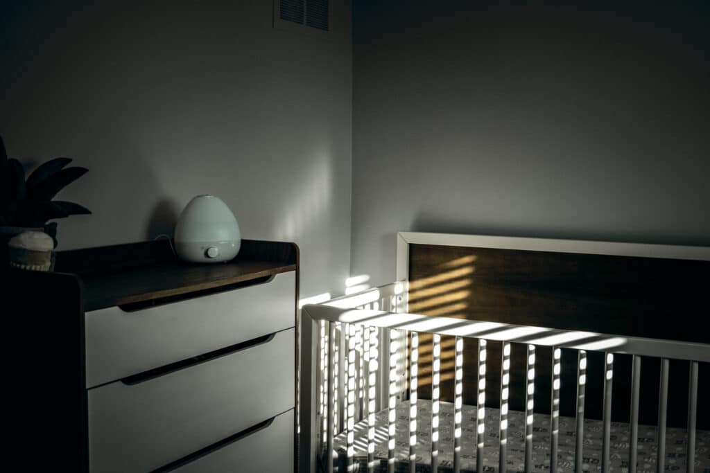 A white crib sits in a corner near a wooden dresser with a small humidifier on top. Sunlight streams through window blinds, casting striped shadows across the crib and the wall.
