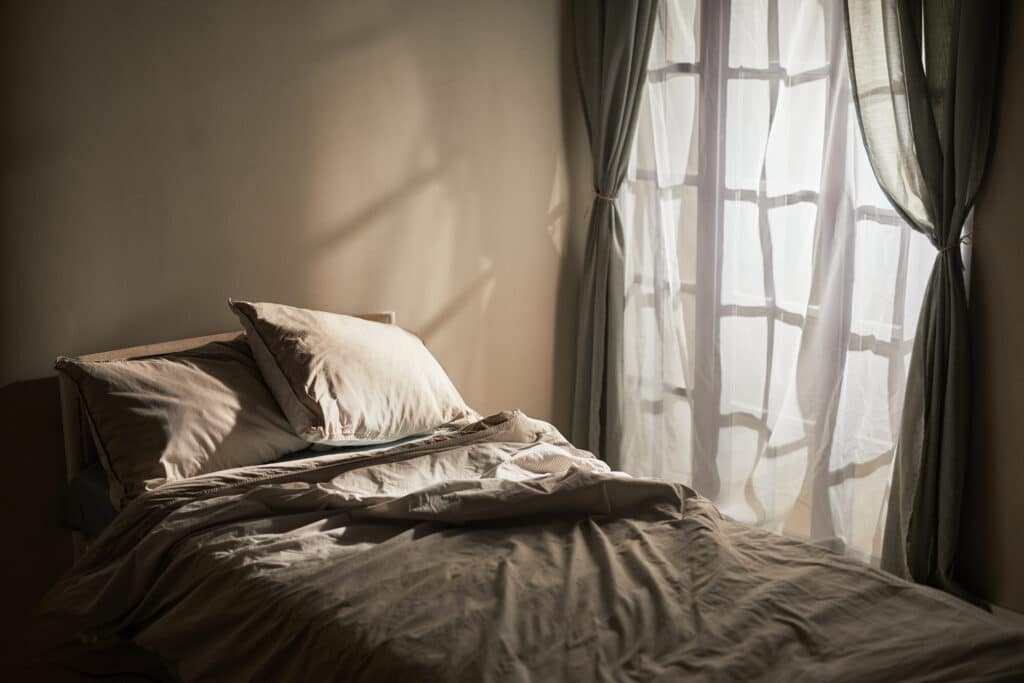 Unmade bed with beige sheets and pillows near a window with sheer curtains. Sunlight casts shadows on the wall and bed, creating a soft, tranquil atmosphere in the room.