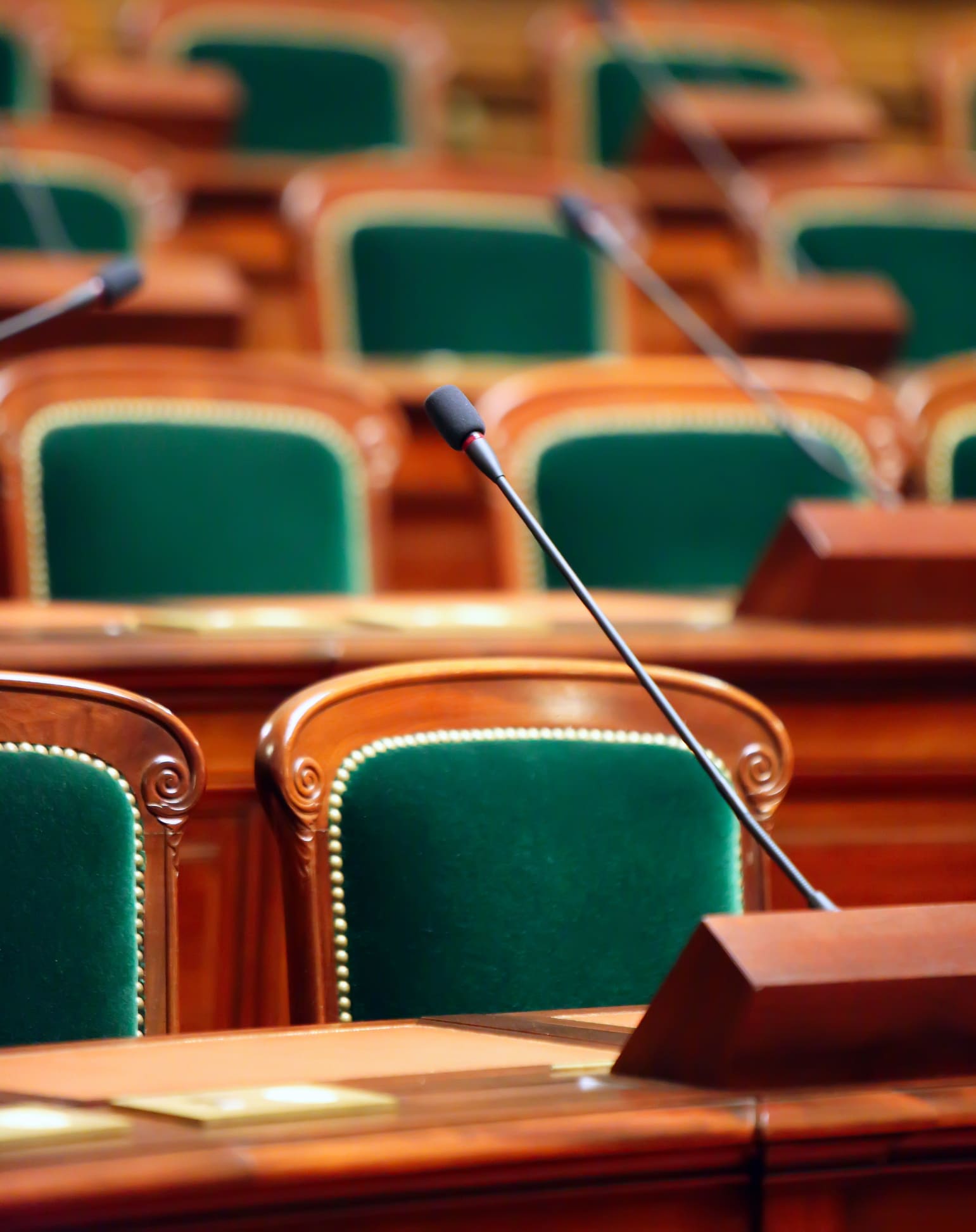 Rows of empty green upholstered chairs with wooden frames and built-in microphones in a legislative or parliamentary chamber, suggesting a formal government setting.