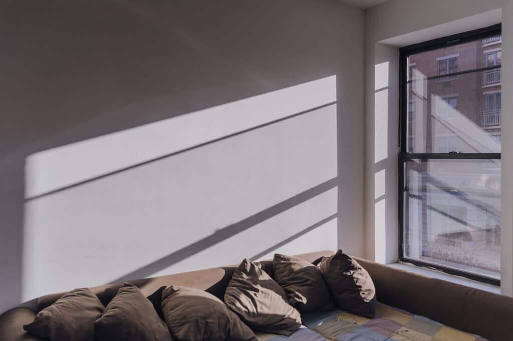 Sunlight casts shadows across a white wall and brown couch with several brown pillows. The window shows part of a brick building outside, and the floor is covered with a colorful patterned blanket or sheet.