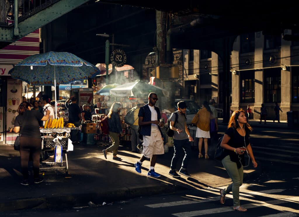 People walk through a busy city street market under an elevated train track. Sunlight creates strong shadows. A vendor sells food under a patterned umbrella. Some people carry drinks or bags.