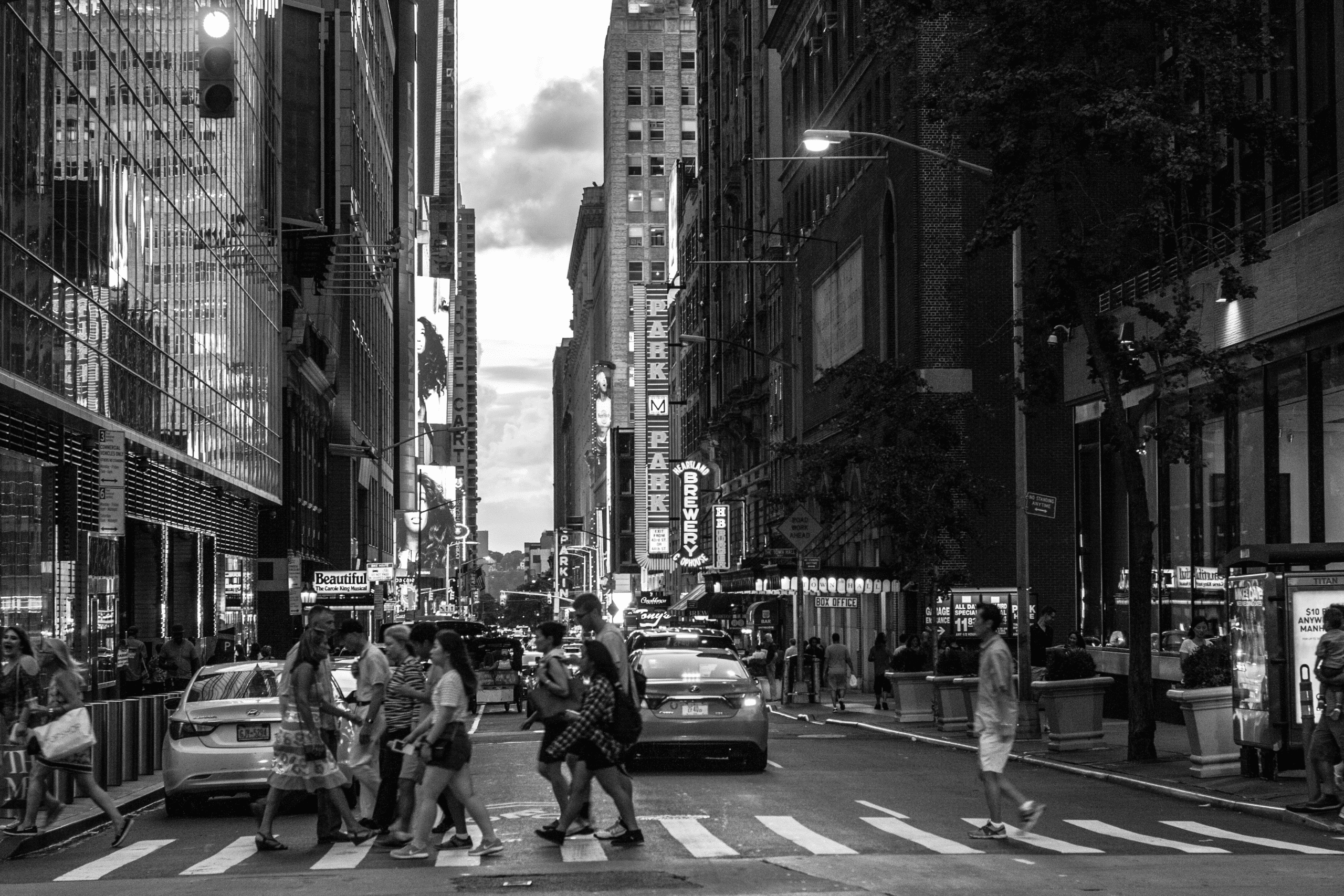 People cross a busy city street at a crosswalk surrounded by tall buildings and parked cars. Neon signs are visible on the buildings. The photo is in black and white.