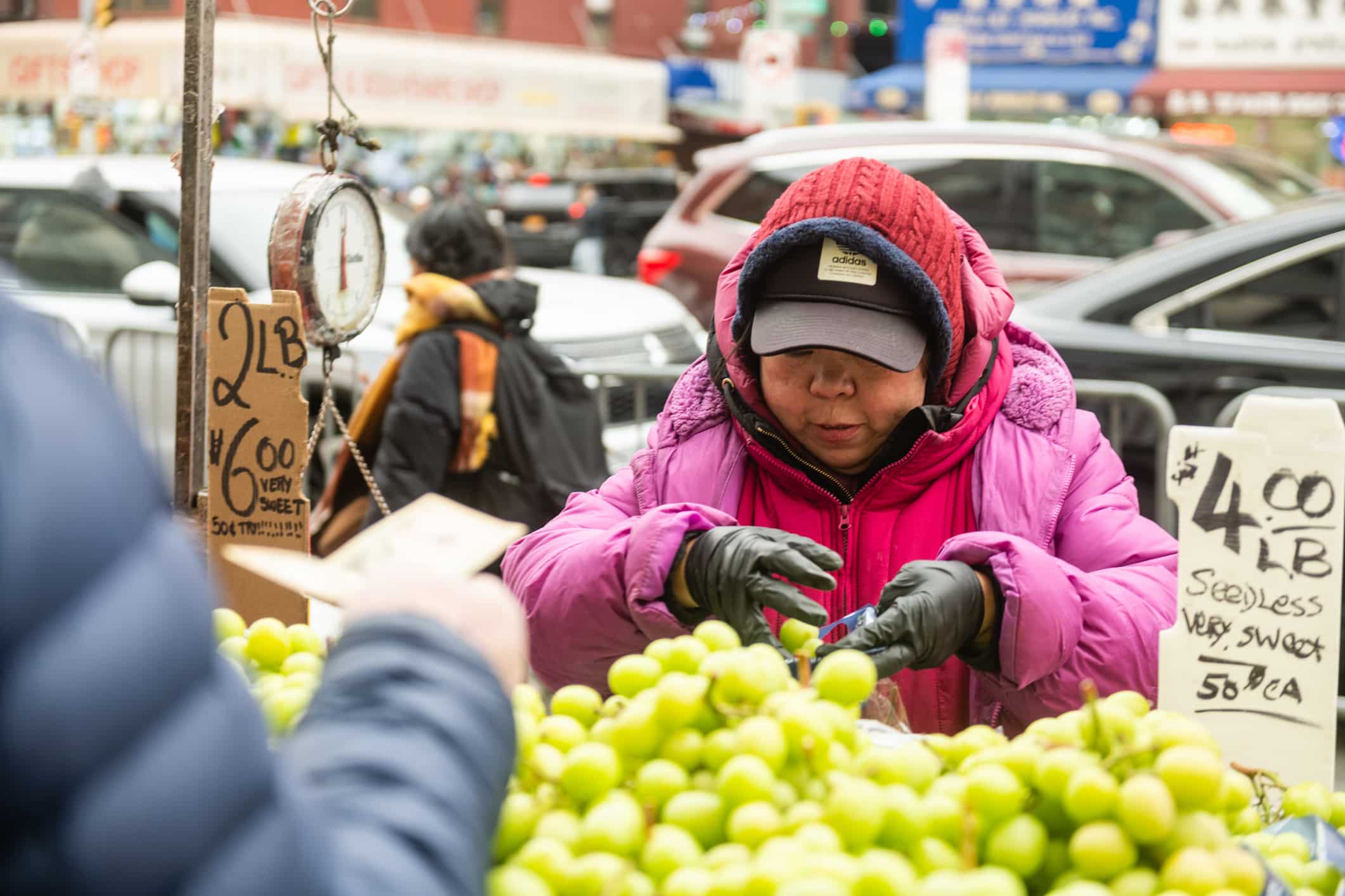 A person in a pink coat and hat selects green grapes at an outdoor market stand. There are price signs, a hanging scale, and cars in the background. Another persons hand holding money is visible in the foreground.