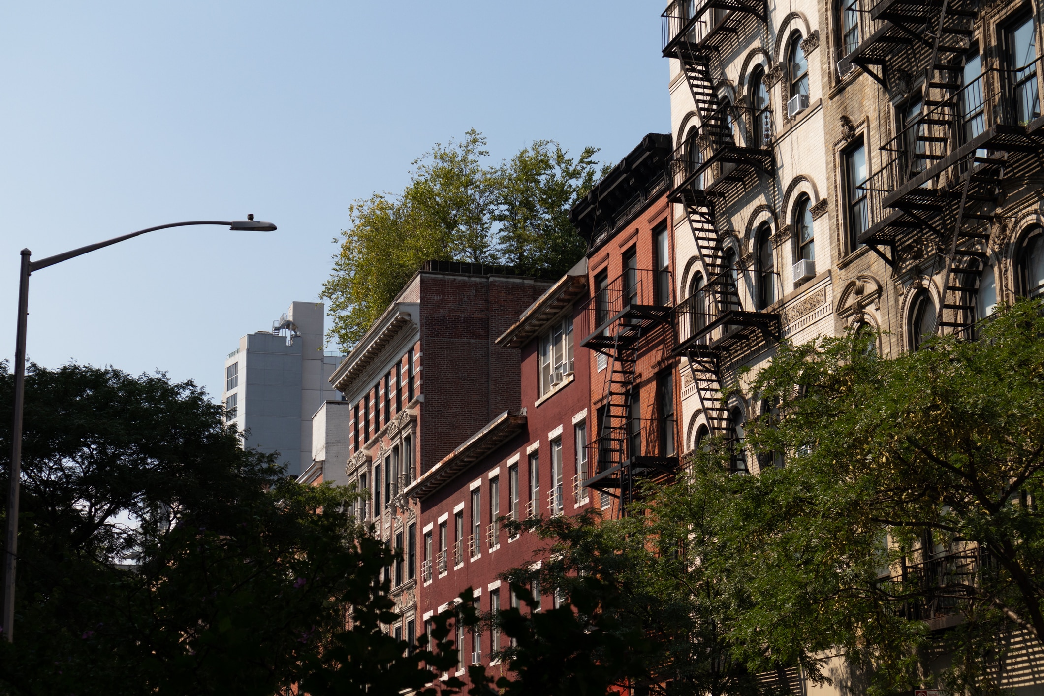 A row of multi-story brick and stone apartment buildings with fire escapes, partially shaded by trees, under a clear blue sky. A streetlight is visible on the left side of the image.