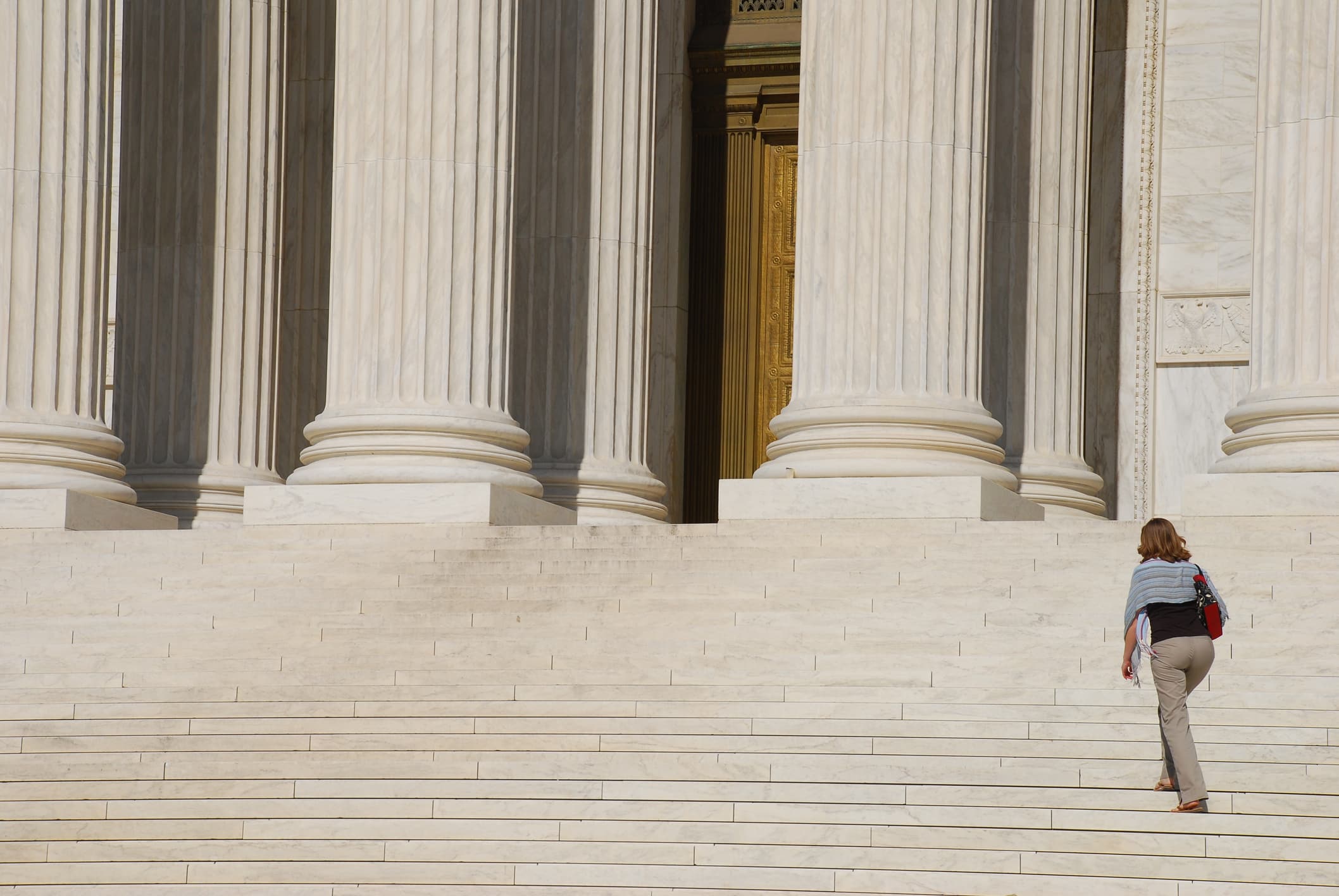 A person walks up wide stone steps toward large columns at the entrance of a grand building, possibly a courthouse or government structure. The scene is mostly empty except for the individual ascending the steps.
