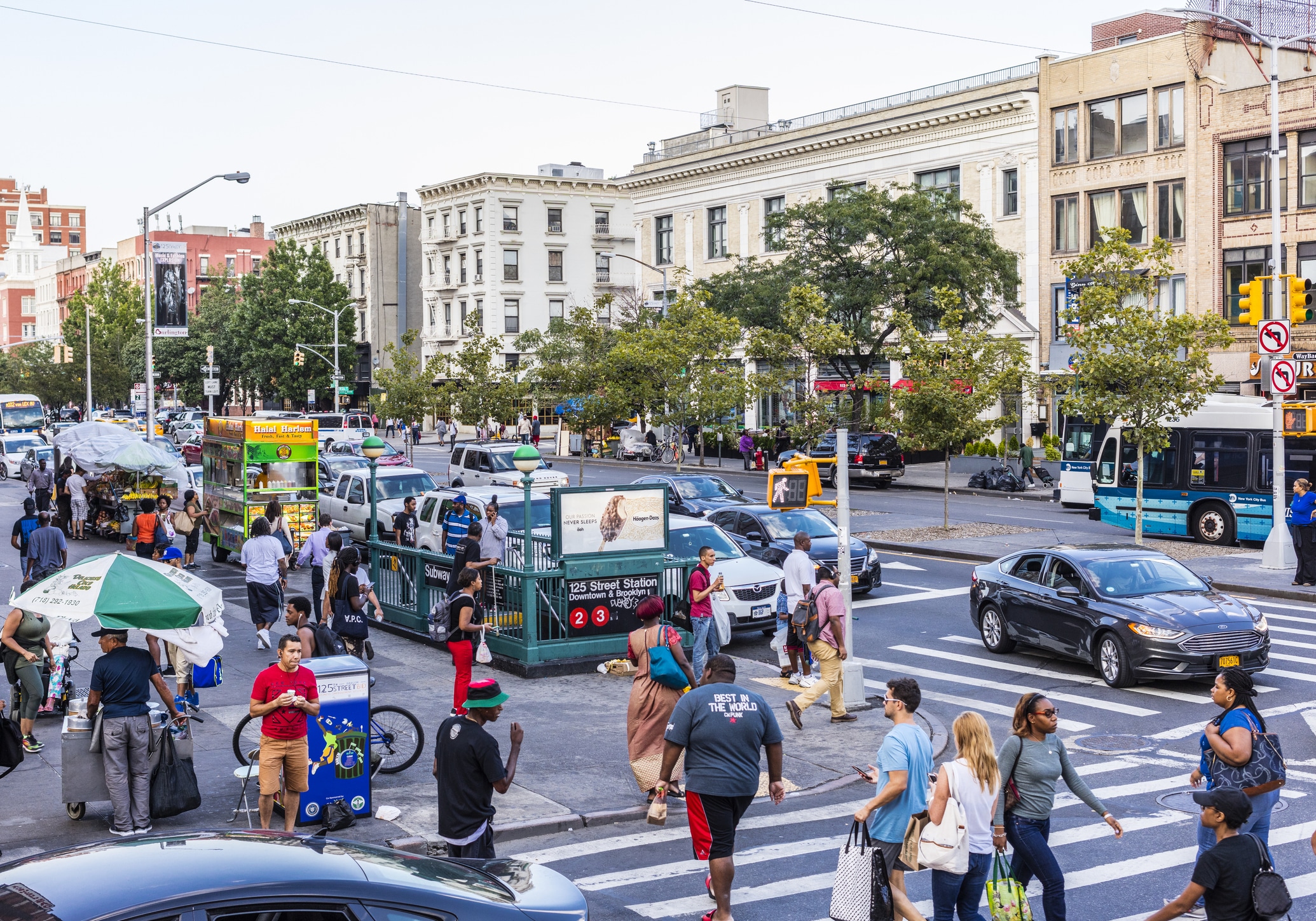 Busy urban street scene with people walking, street vendors, cars, and buildings in the background. A subway entrance and traffic signals are visible, along with trees lining the sidewalk. Urban activity is prominent.