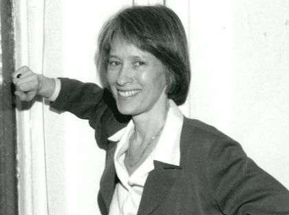 A woman with short hair smiles at the camera. She is dressed in a blazer over a blouse and is standing indoors, with one arm bent and resting against a surface. The image is in black and white.