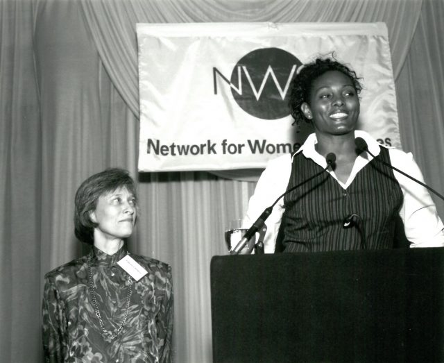 Two women stand at a podium with microphones. One is speaking while the other looks on. A banner behind them reads Network for Women with part of the text obscured. The setting appears to be a formal event or conference.