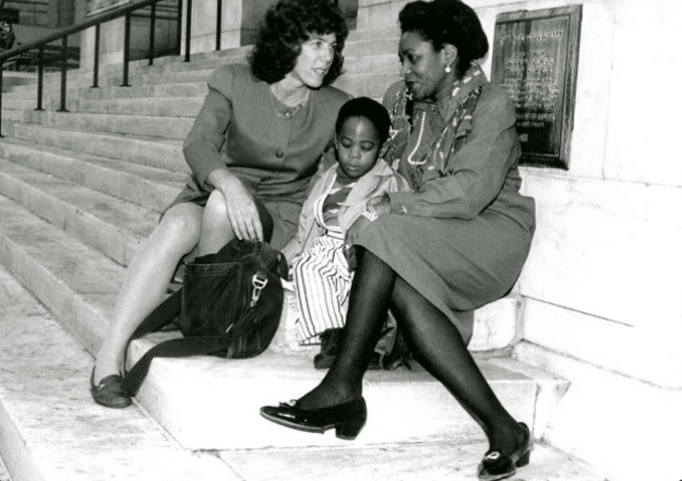 Two women sit on stone steps, facing each other, with a young girl between them. The women appear to be in conversation, while the girl looks down. All are dressed in formal attire. A large bag sits near one woman.