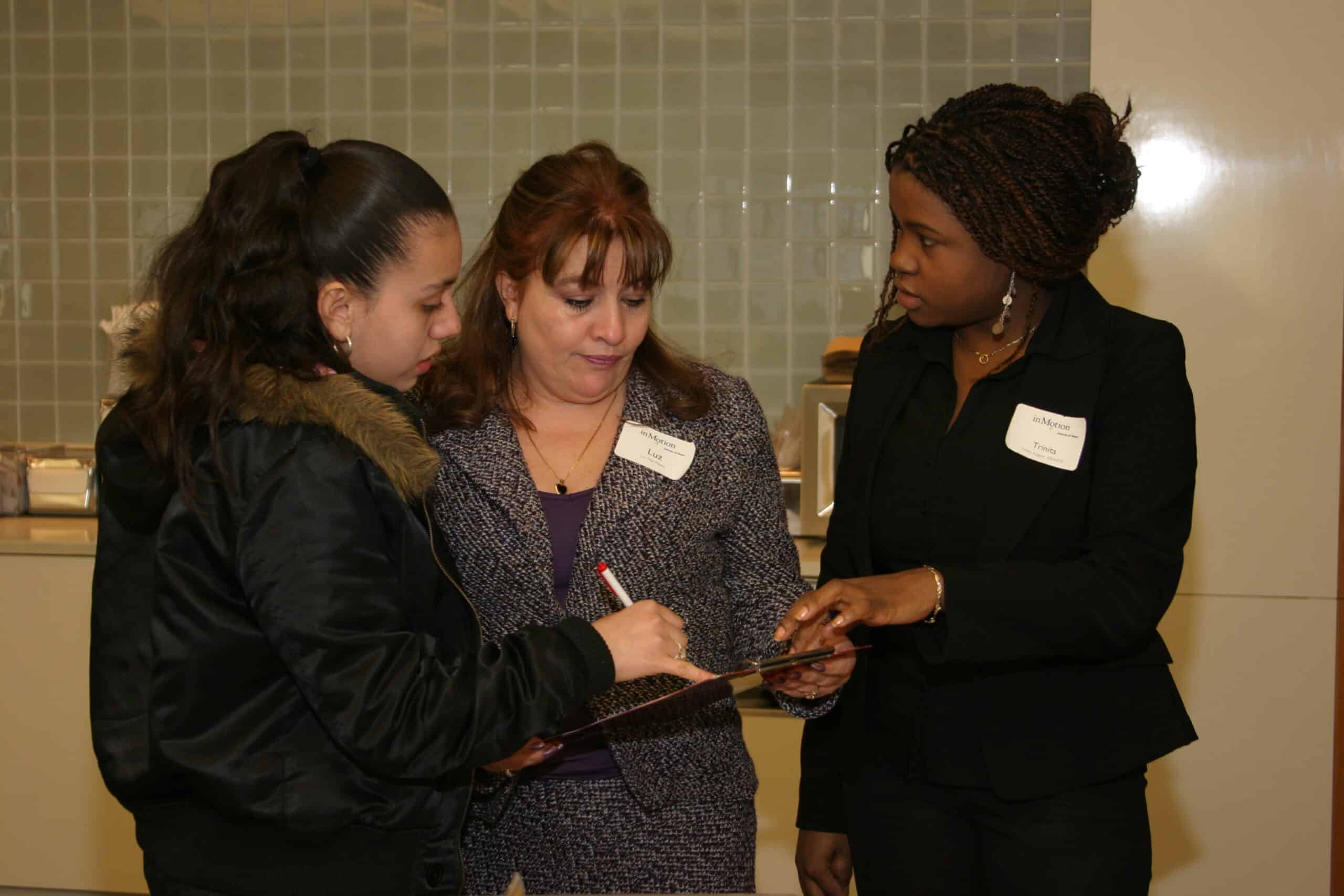 Three women stand indoors, one holding a pen and clipboard while another points to it. All wear name tags and are engaged in conversation. The background features a tiled wall and countertop.