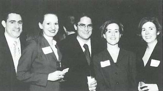 Five people in business attire stand close together at an indoor event, each wearing name tags. Two hold drinks. They face the camera and smile. The photo is in black and white.