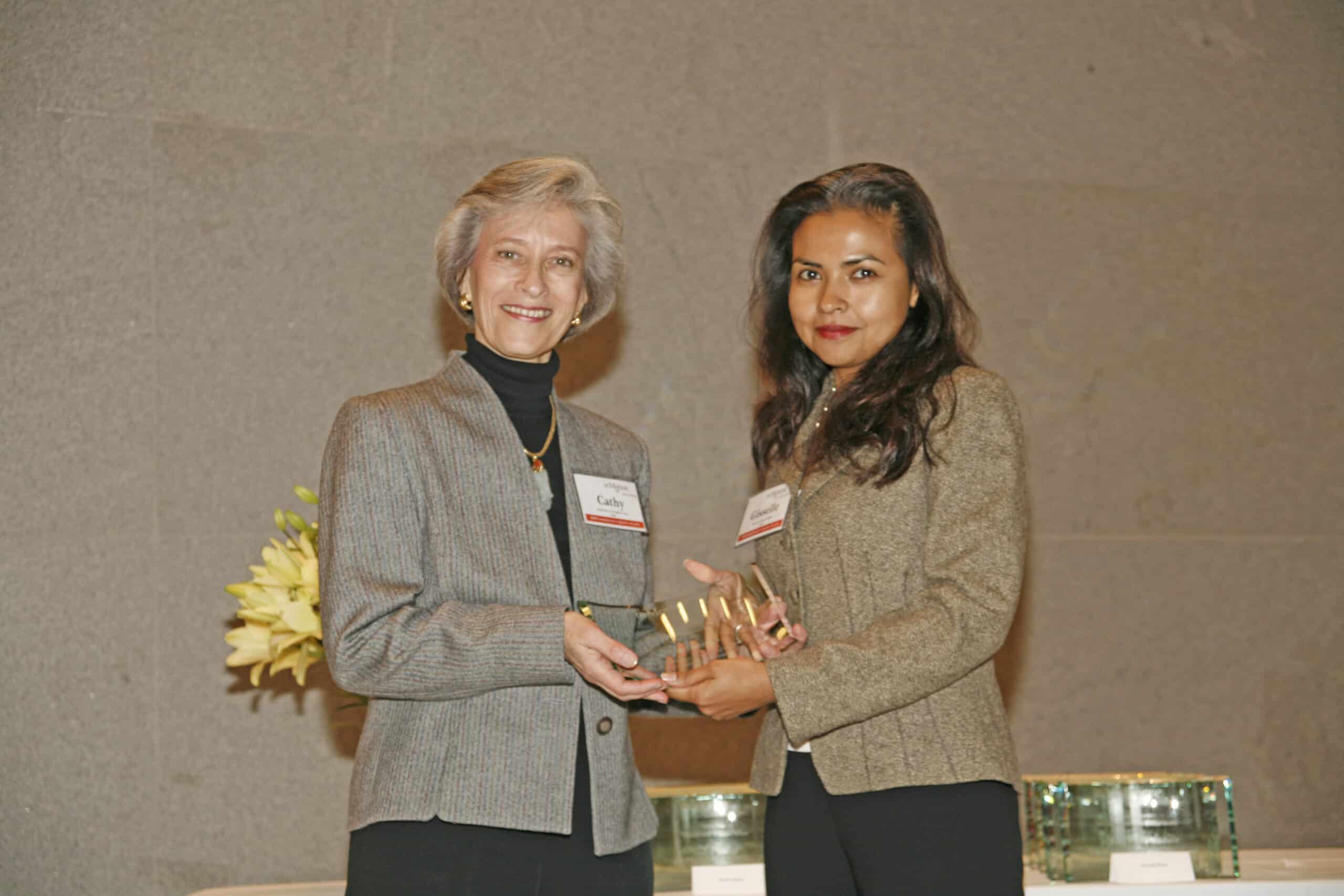 Two women in business attire stand side by side, holding an award together. Both are facing the camera and smiling. There are glass awards and a bouquet of yellow flowers on a table in the background.
