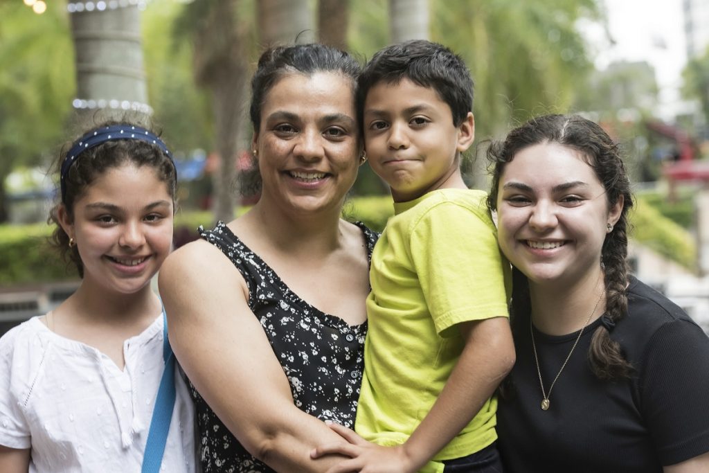 A woman stands outdoors, smiling, with three children—two girls and a boy. The boy is being held in her arms. Trees and greenery are in the background. All four are looking at the camera.