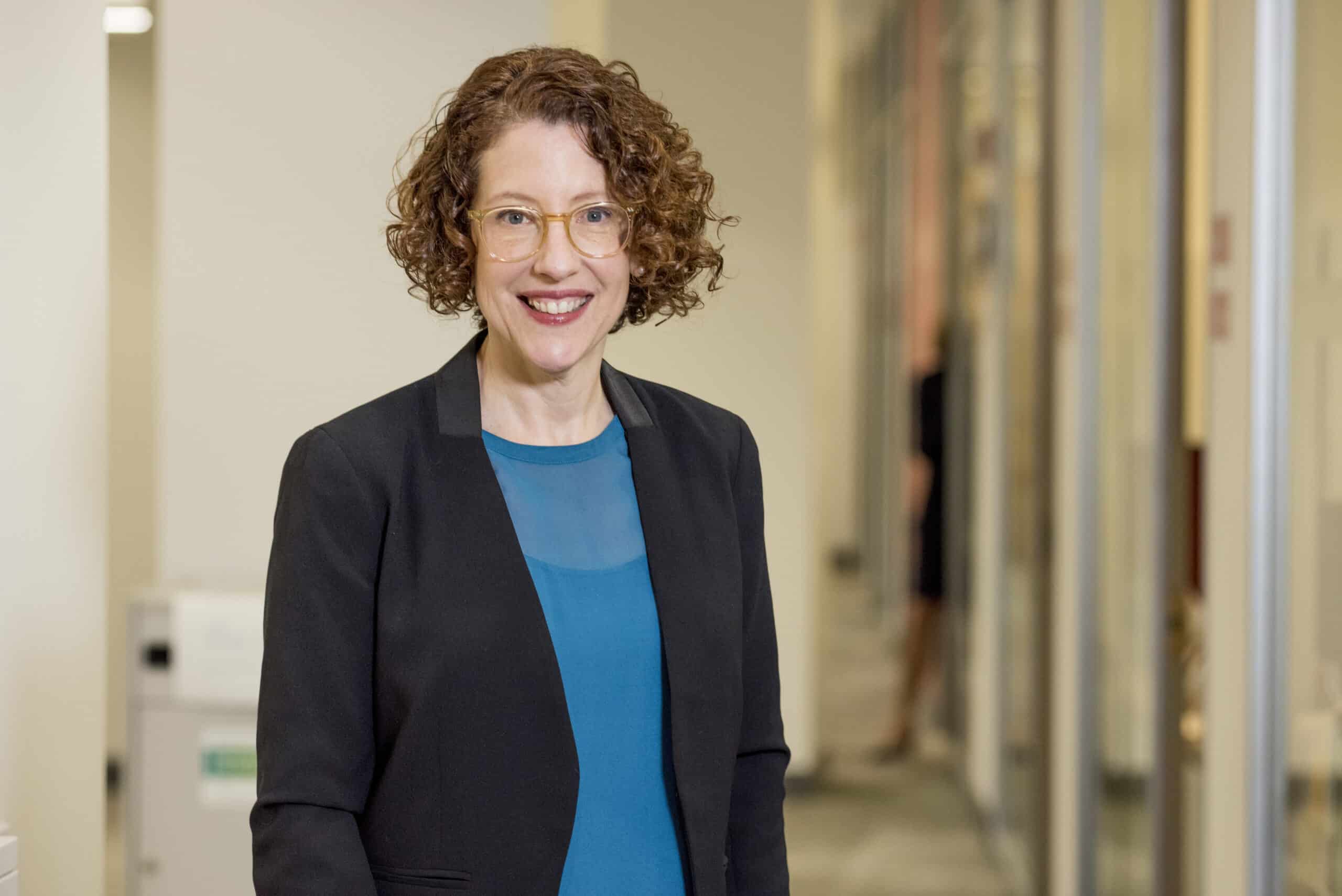 A woman with curly hair and glasses is smiling, wearing a teal top and black blazer, standing in a hallway with glass doors and blurred figures in the background.