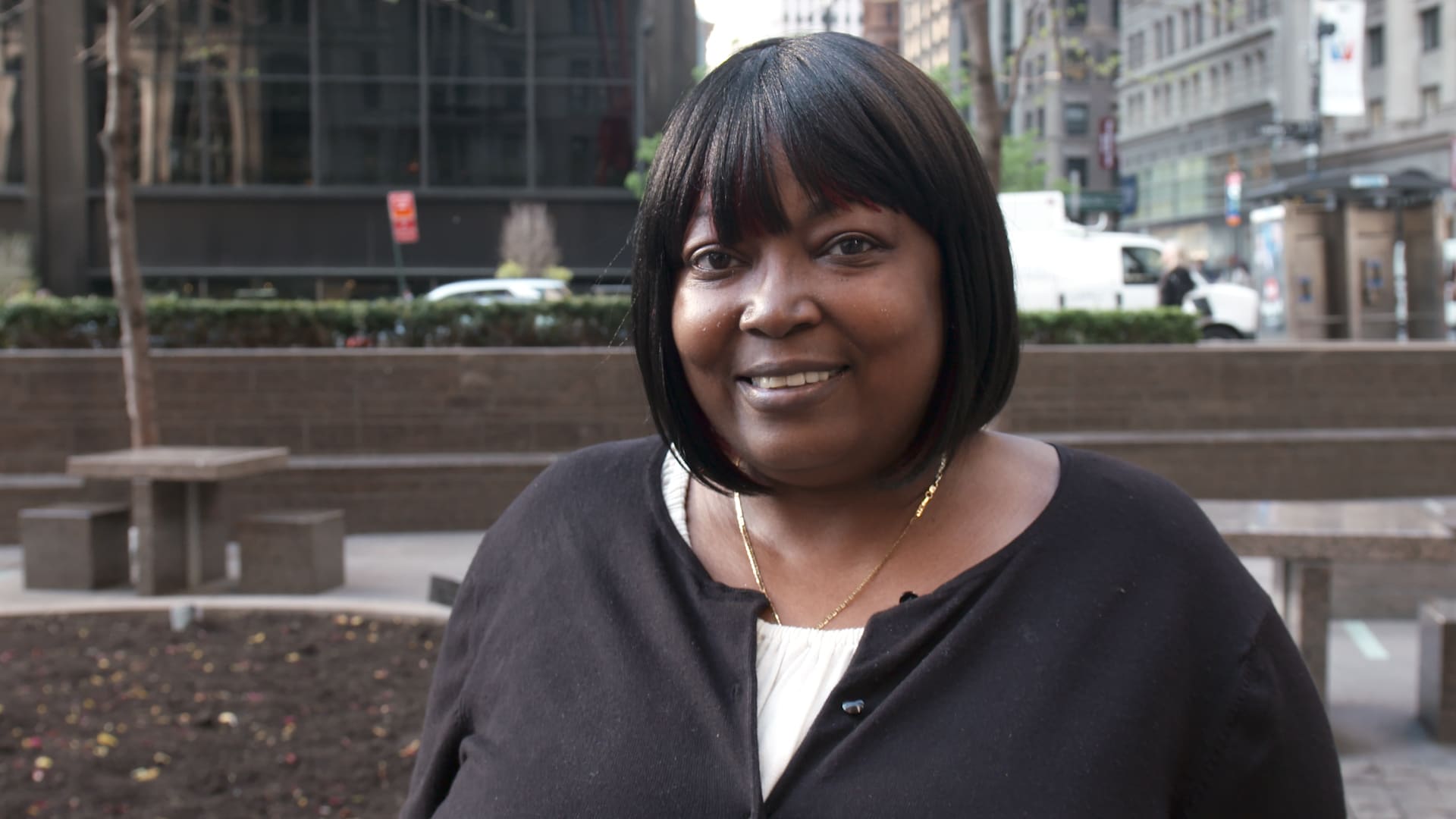 A woman with straight black hair and a dark cardigan stands outdoors in an urban setting, smiling at the camera. Buildings, cars, and trees are visible in the background.