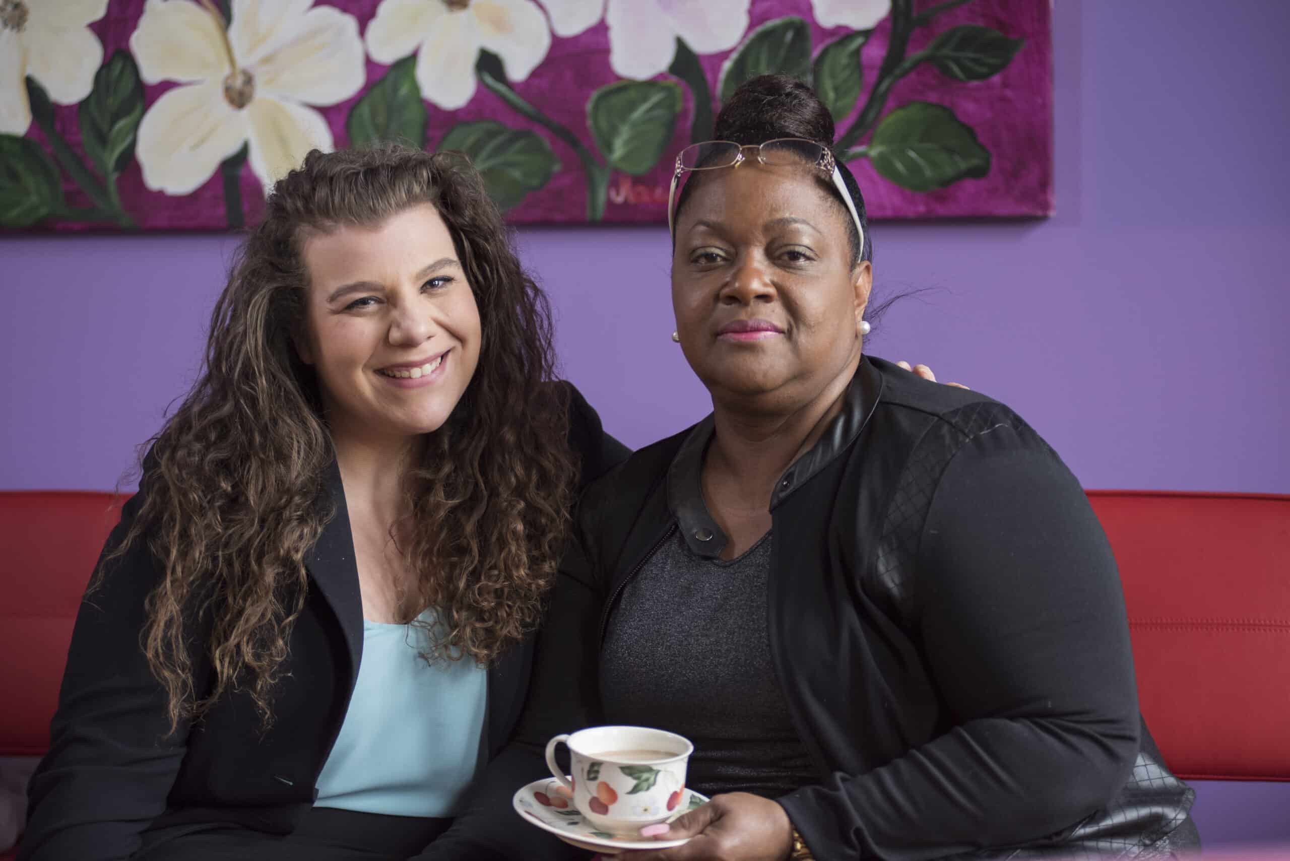 Two women sit together on a red couch in front of a purple wall with floral art. One woman smiles with her arm around the other, who holds a teacup and saucer. Both face the camera.