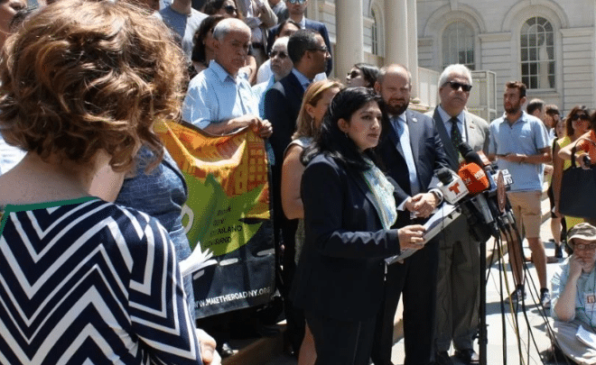 A woman speaks at a podium with multiple microphones outside a government building, surrounded by a crowd of people standing and listening. Some hold banners. Journalists are present with cameras and recording devices.