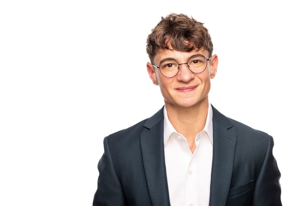 A person with short brown hair, glasses, and a dark suit jacket over a white collared shirt stands against a plain white background, facing the camera with a slight smile.