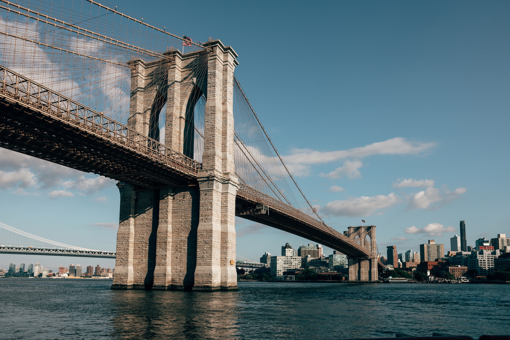 The Brooklyn Bridge spans the East River in New York City, connecting Manhattan and Brooklyn. The bridges stone towers and steel cables are visible, with city buildings and a partly cloudy sky in the background.