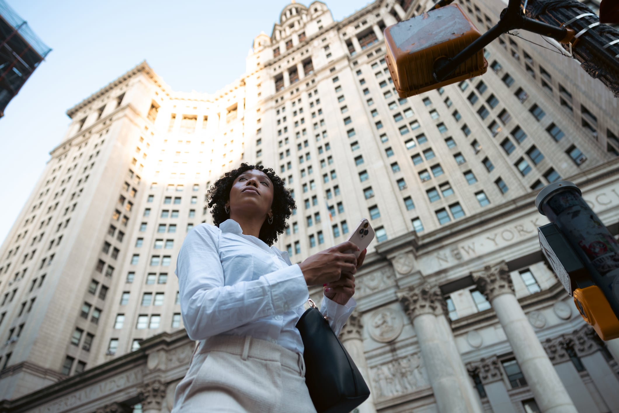 A person in a white shirt holds a smartphone and stands in front of a tall, ornate stone building under daylight. The photo is taken from a low angle, emphasizing the height of the building.