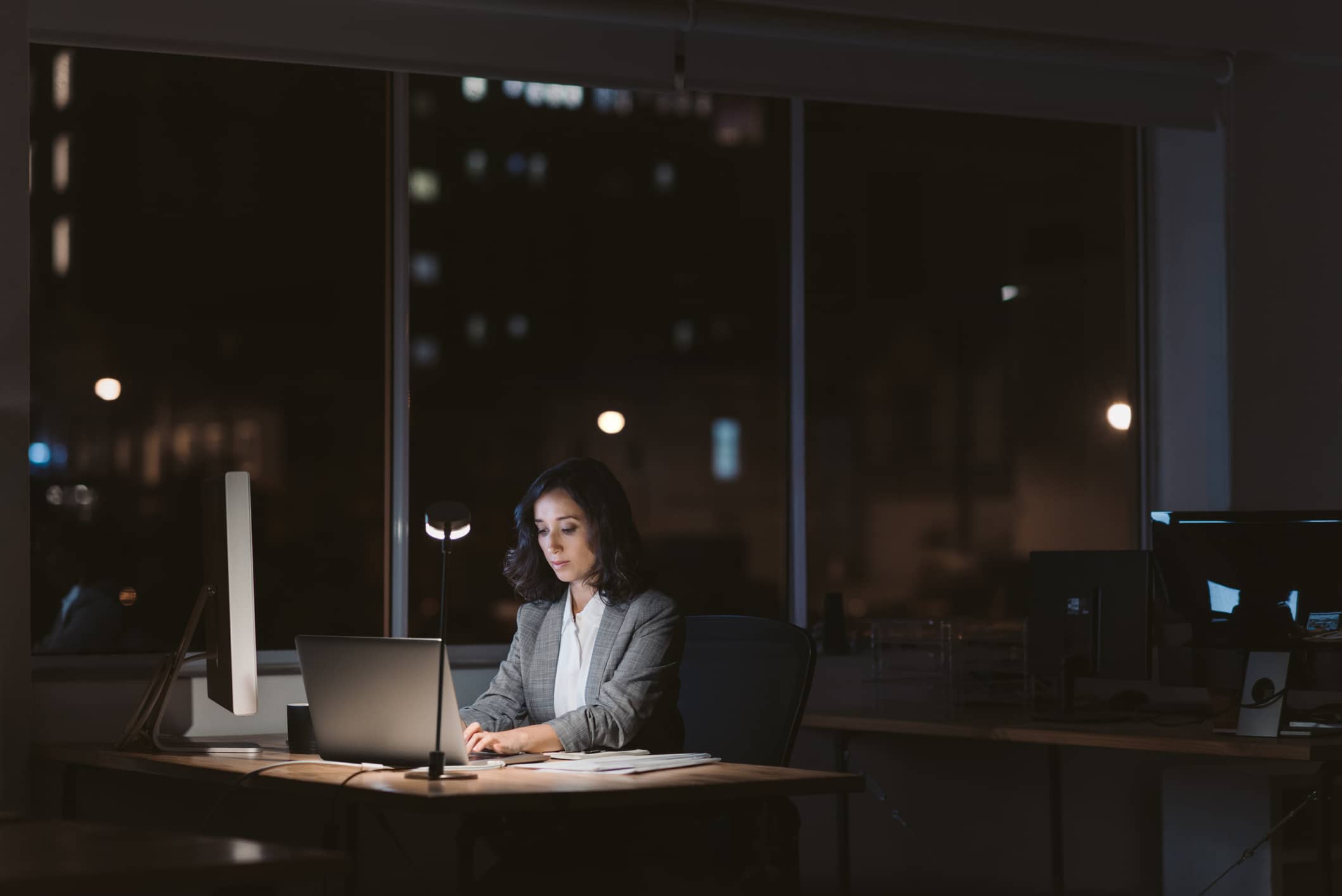 A woman in business attire works on a laptop at a desk in a dark office at night, illuminated by a desk lamp. Large windows behind her show city lights.