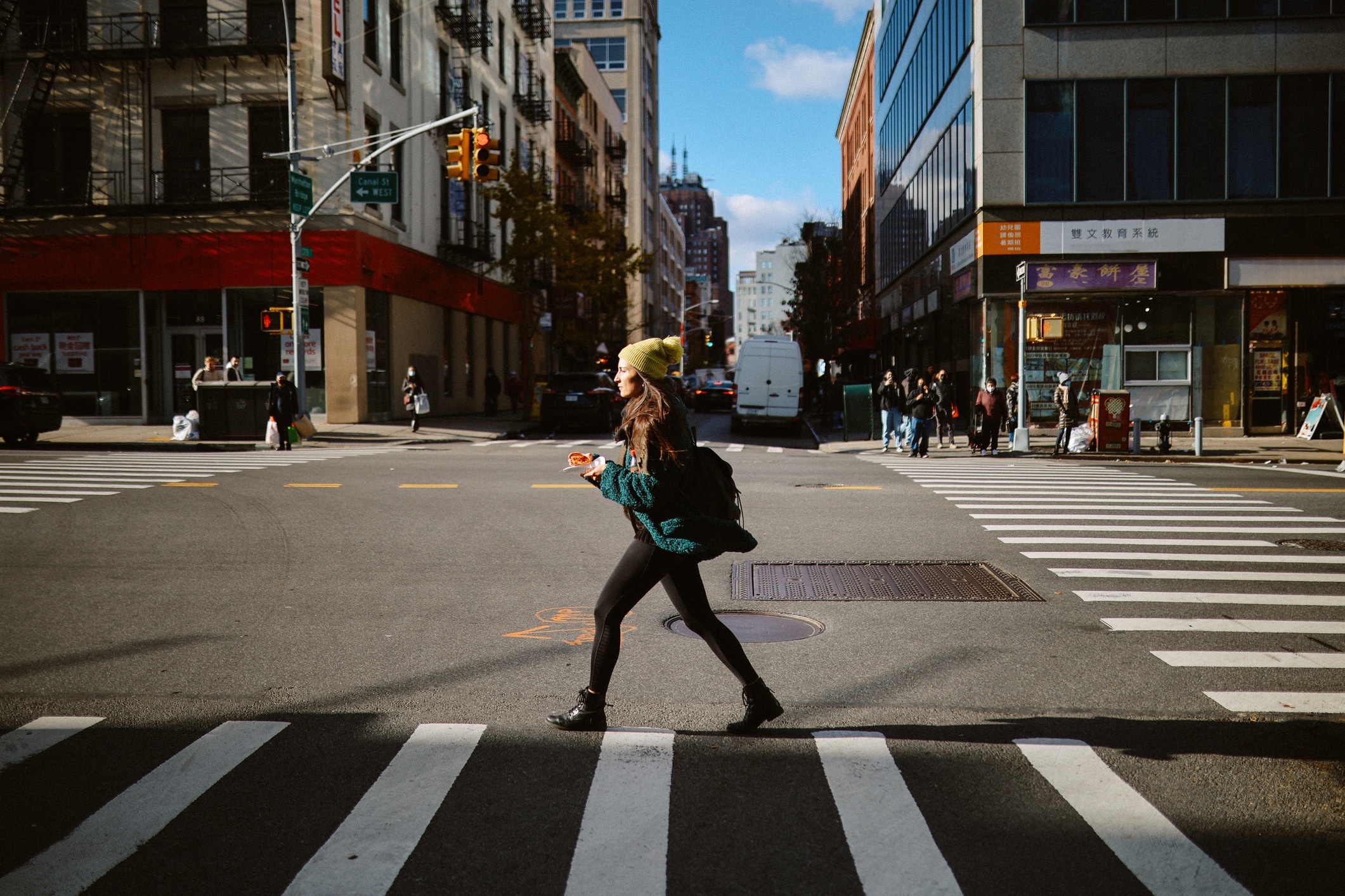A person wearing a yellow hat and dark clothing walks across a city street crosswalk holding a drink. Buildings, pedestrians, parked cars, and traffic lights are visible in the background under a bright sky.
