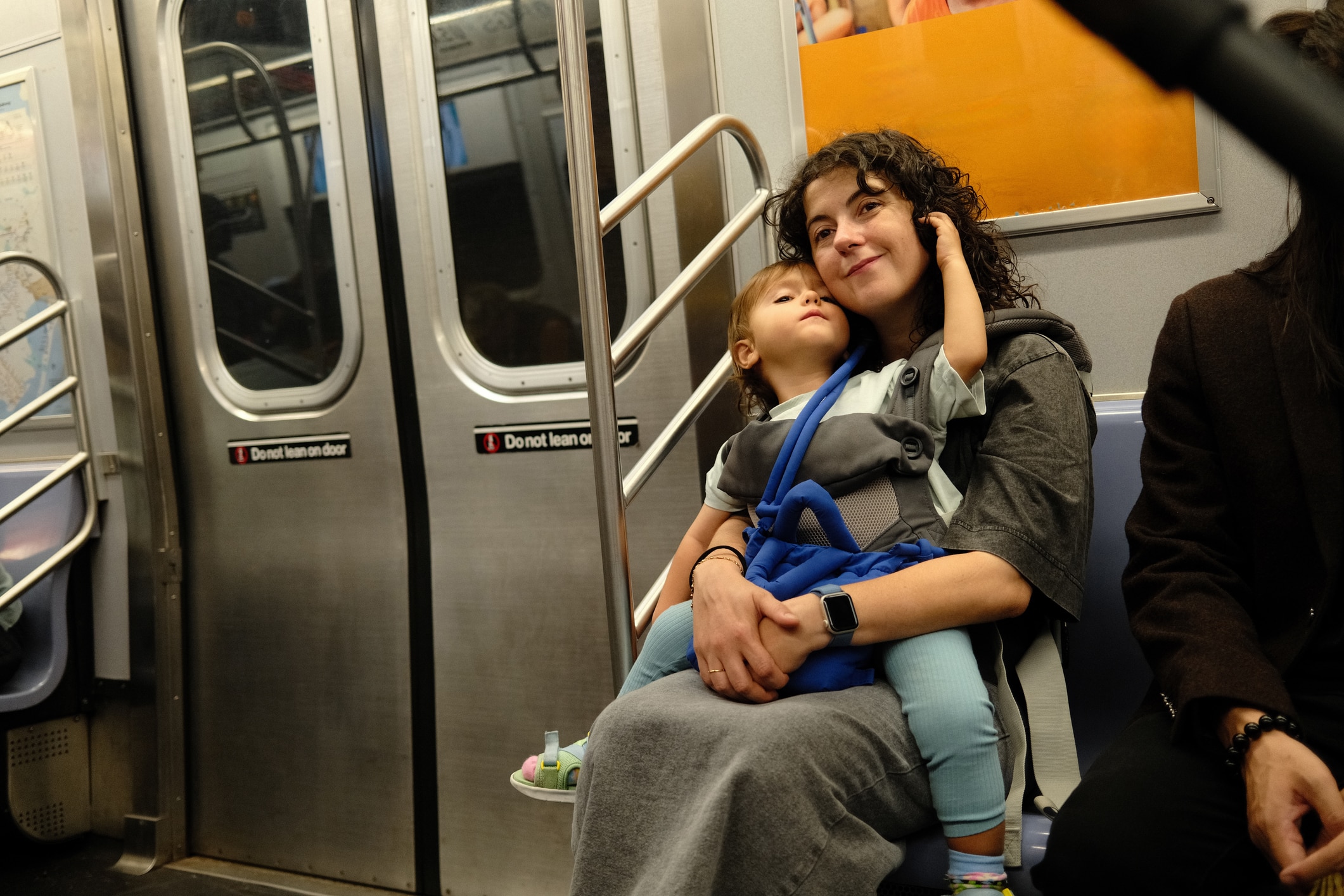 A woman sits on a subway bench, holding a small child in her lap. Both appear relaxed. The subway doors behind them have warning signs, and another person is partially visible to the right.