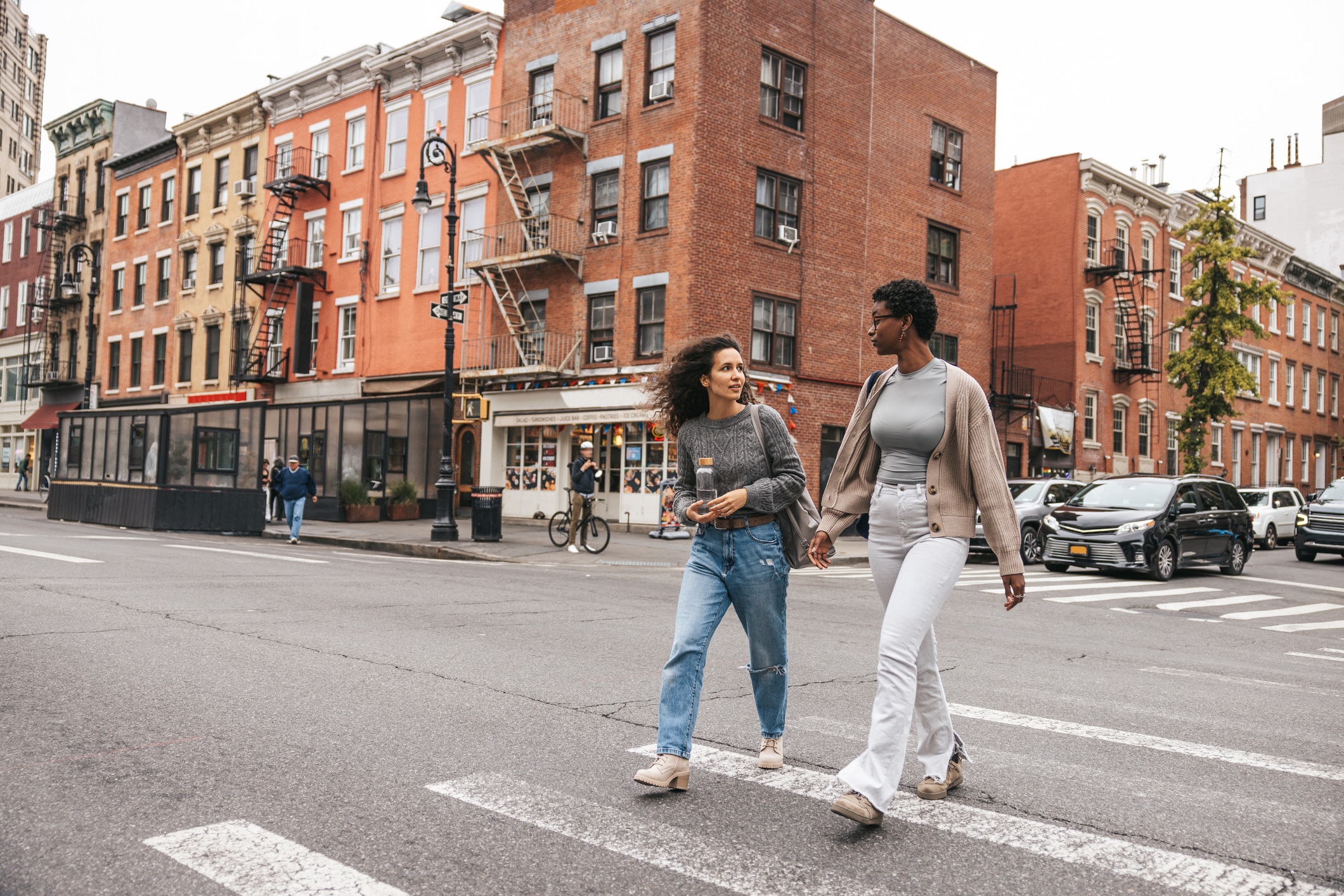 Two people walk across a city street crosswalk, talking and holding drinks. Brick apartment buildings, parked cars, and a few pedestrians are visible in the background on an overcast day.