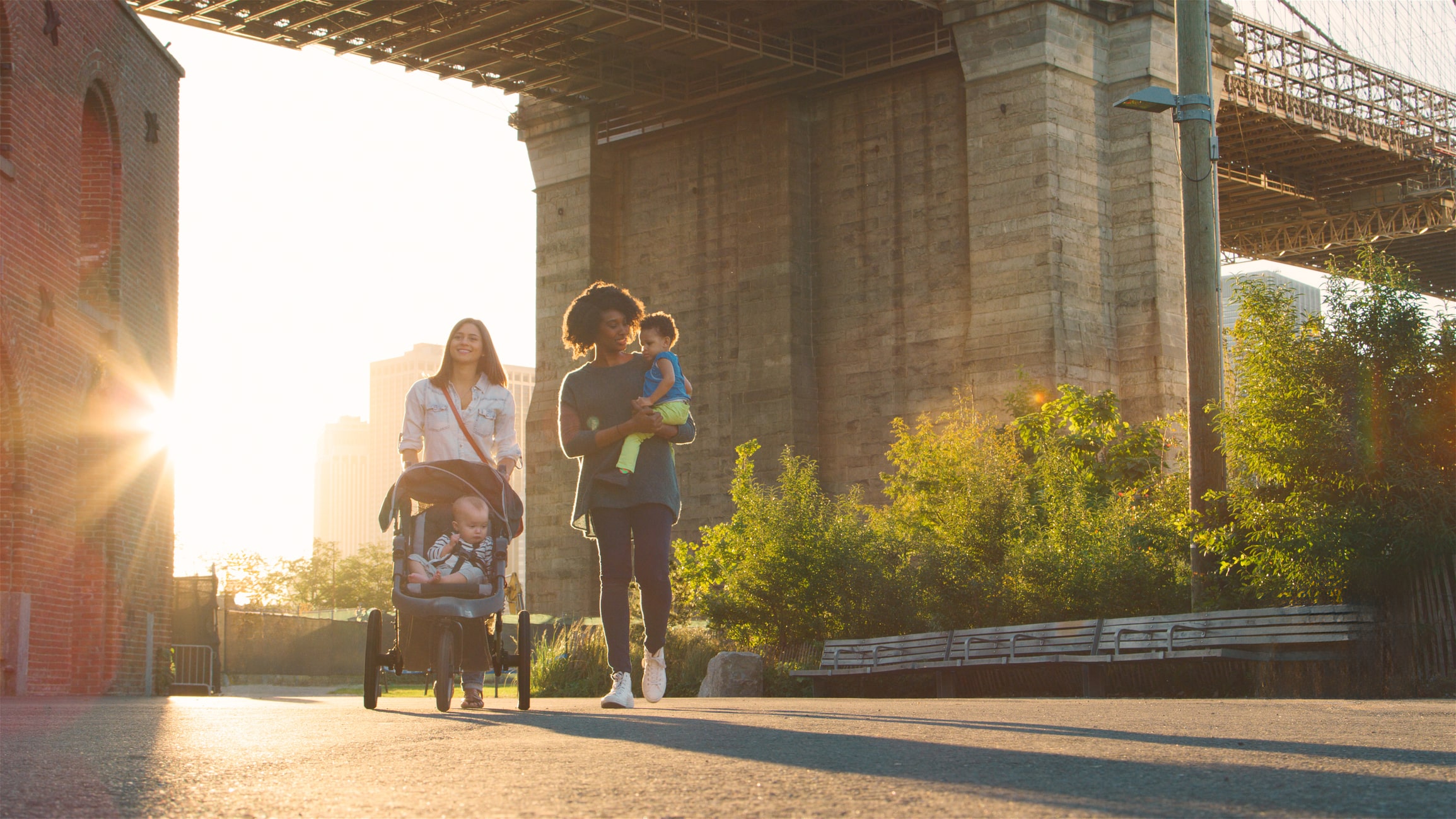 Two adults walk outdoors with a stroller and a young child, passing under a large bridge in a city. Sunlight streams through the trees and buildings in the background.
