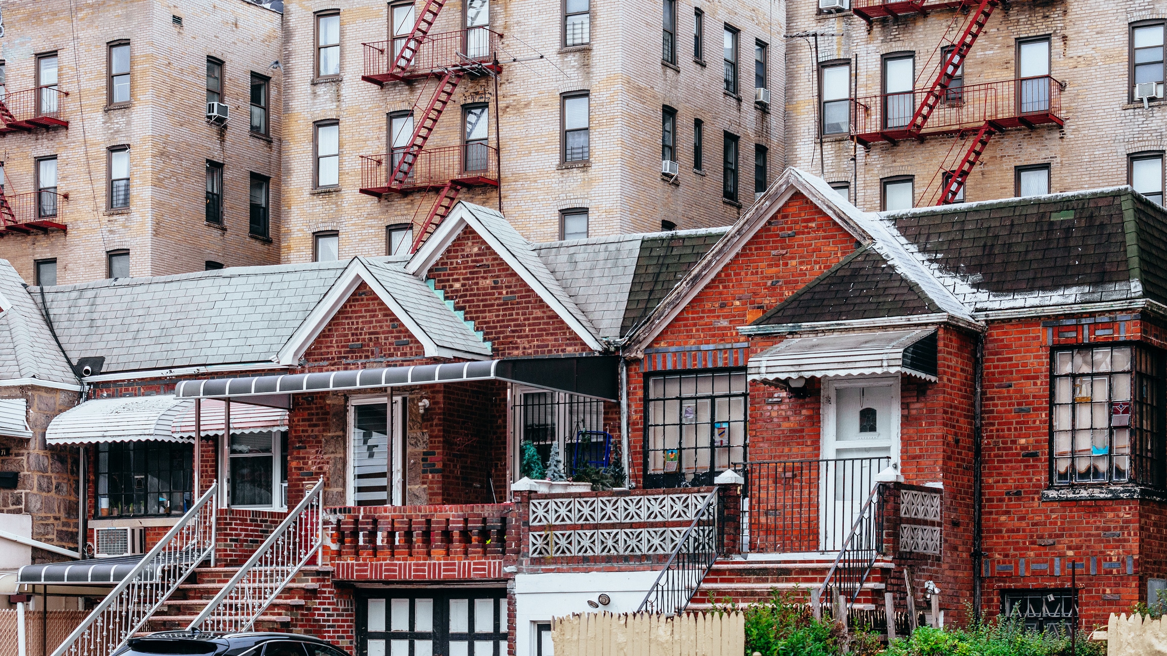 Row of brick houses with small porches and stairs in the foreground, with taller apartment buildings featuring fire escapes in the background. The buildings appear closely packed in an urban neighborhood.