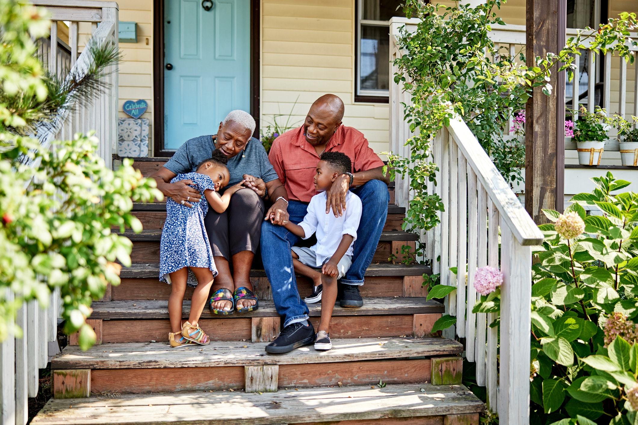 Two adults and two children sit on wooden steps outside a house. One child, wearing a blue dress, leans on an older adult. The other child and adult sit beside them, smiling and holding hands. Plants and flowers surround the porch.