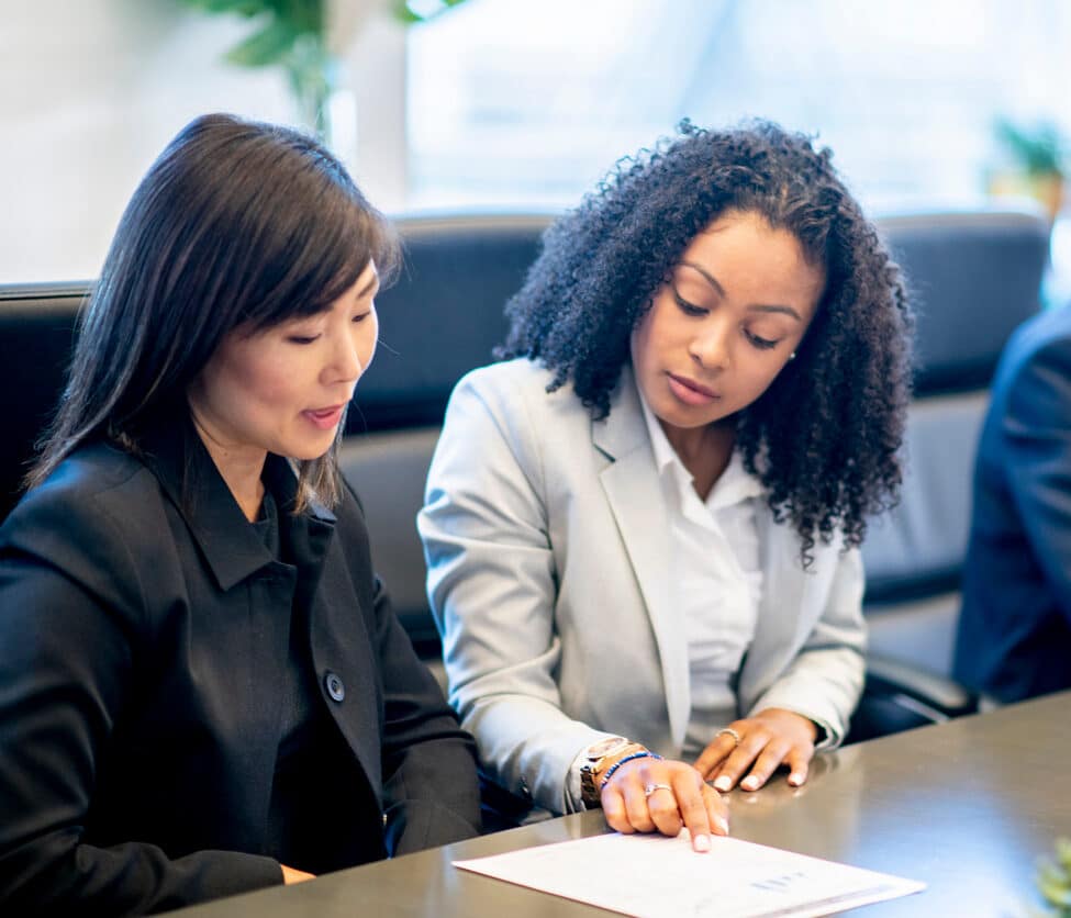 Three people sit at a conference table. Two women in business attire review a document together, while a man in a suit sits nearby, looking away from them.