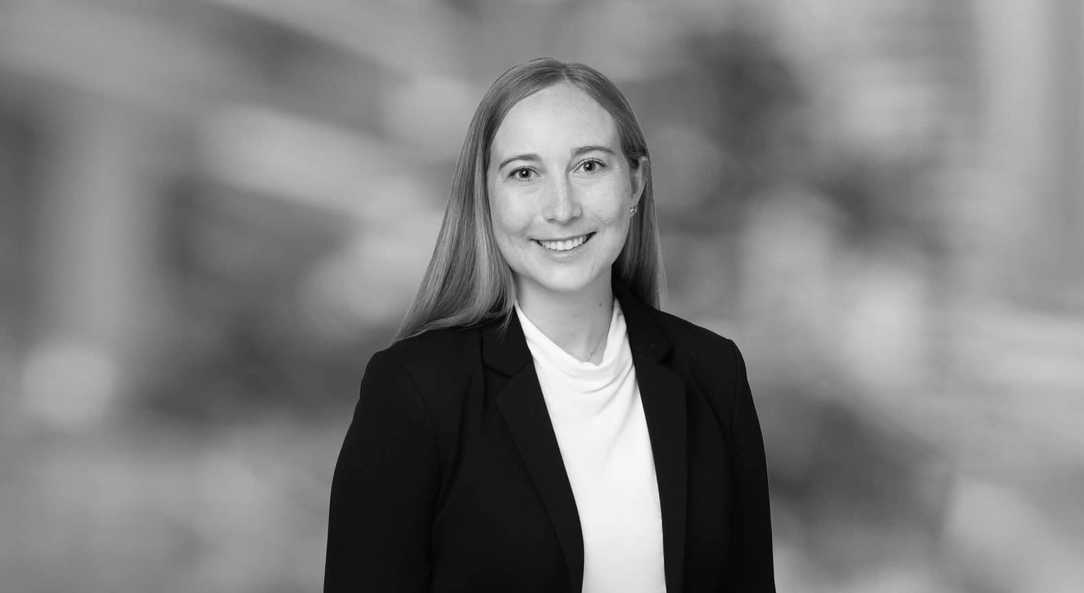 A woman with long straight hair wearing a dark blazer and light turtleneck smiles at the camera. The background is blurred, showing an urban or office environment in grayscale.