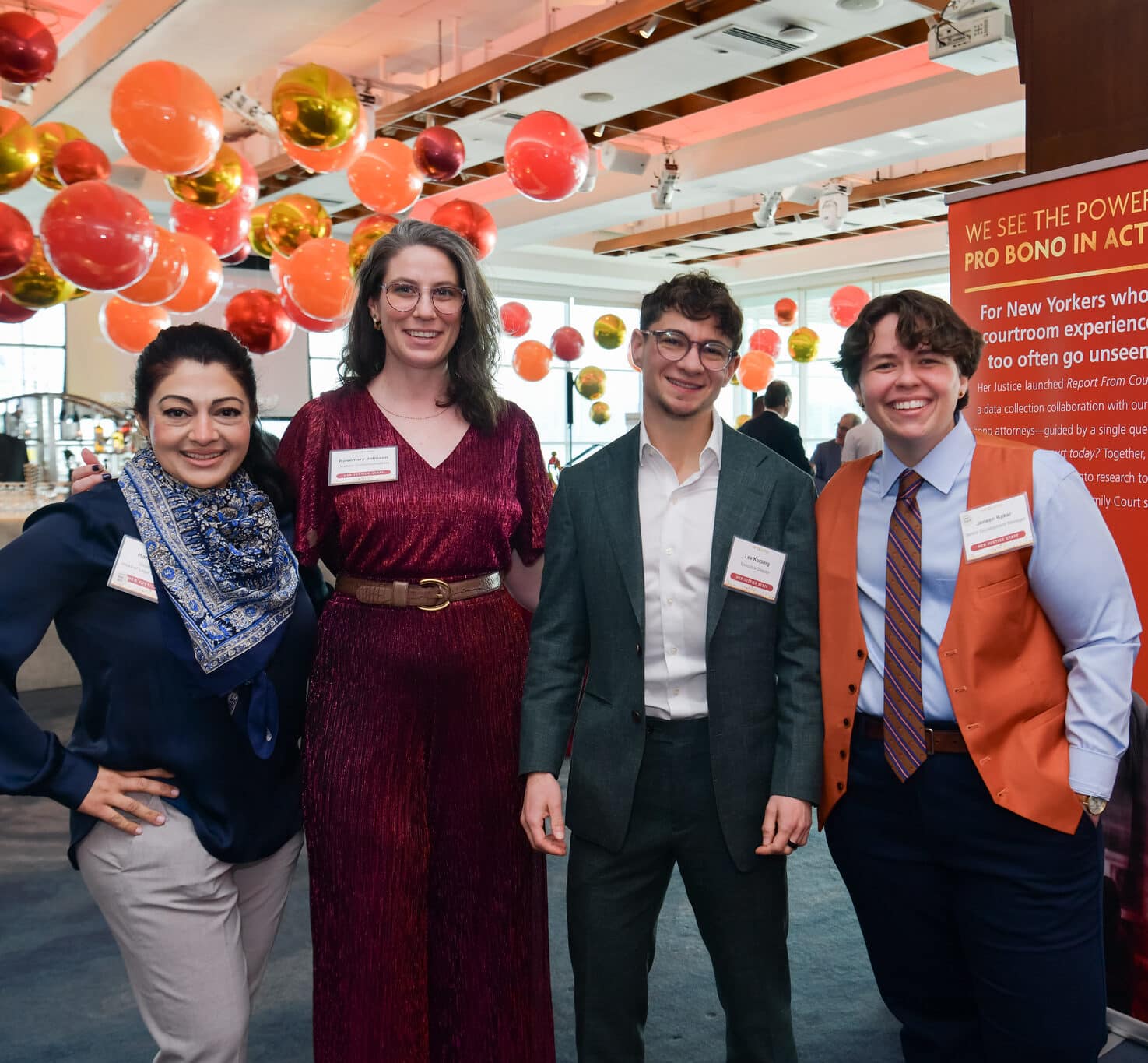 Five people stand together at an indoor event with orange, red, and gold balloons overhead. They wear business or semi-formal attire and name tags. Event signs with text are in the background.