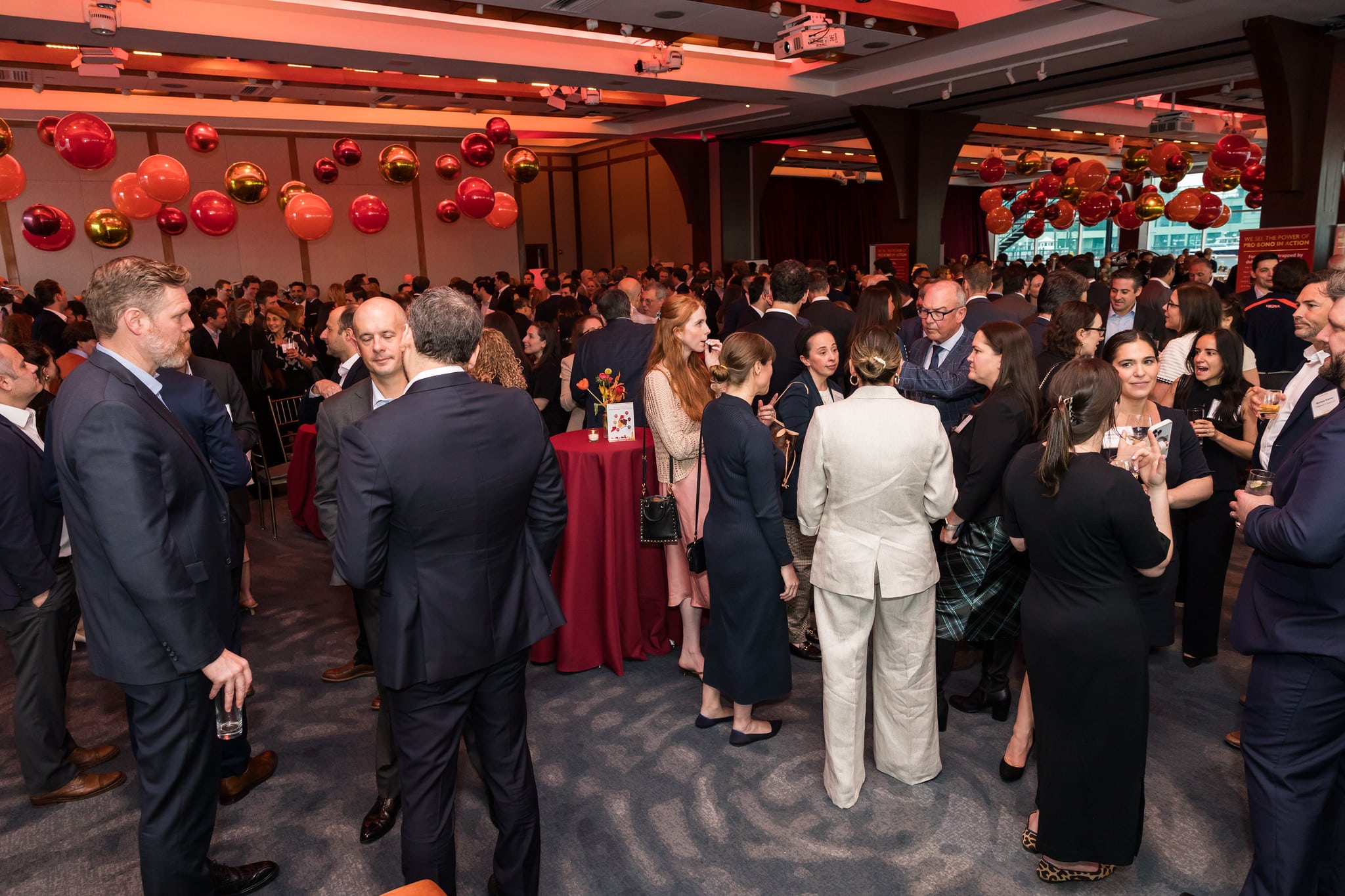A large group of people in formal attire are mingling at an indoor event with red tablecloths and red and orange balloons hanging from the ceiling.