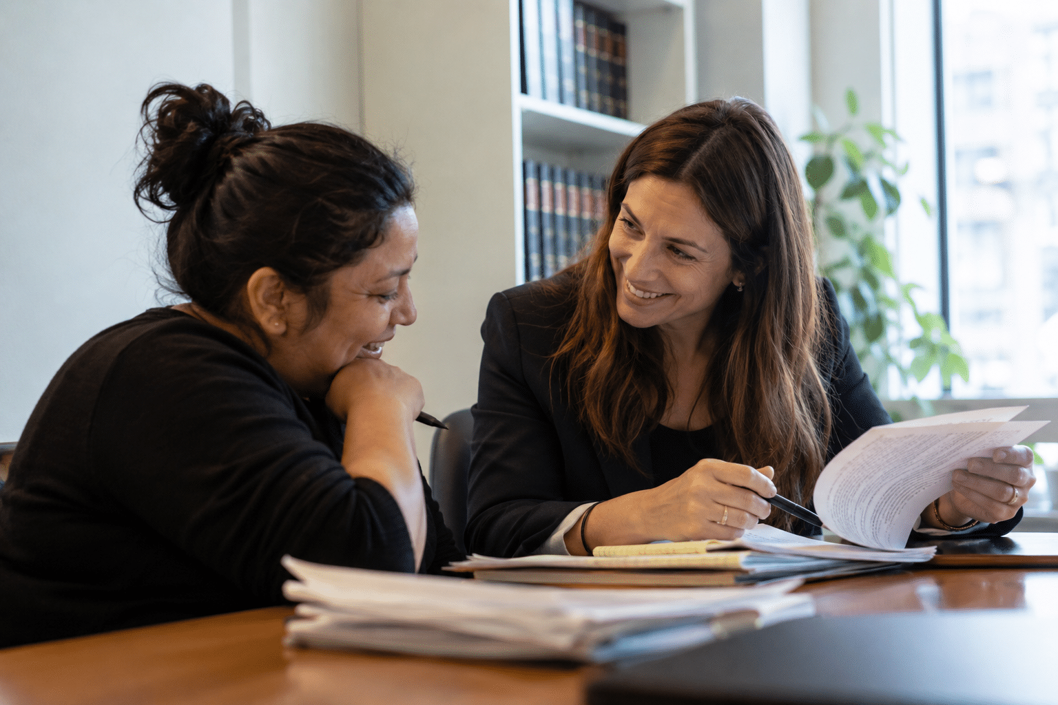 Two women sit at a table covered with papers and folders, talking and smiling. One woman holds documents and a pen, while the other listens, both appearing engaged in a professional office setting.
