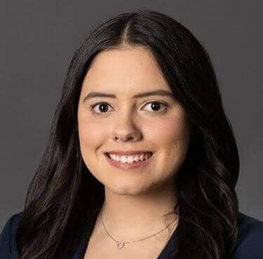 A woman with long, dark hair wearing a navy blazer and white top smiles at the camera against a plain gray background.