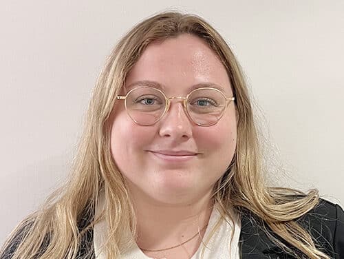 A person with long blonde hair and glasses is wearing a white shirt and black blazer, standing in front of a plain white wall and smiling at the camera.