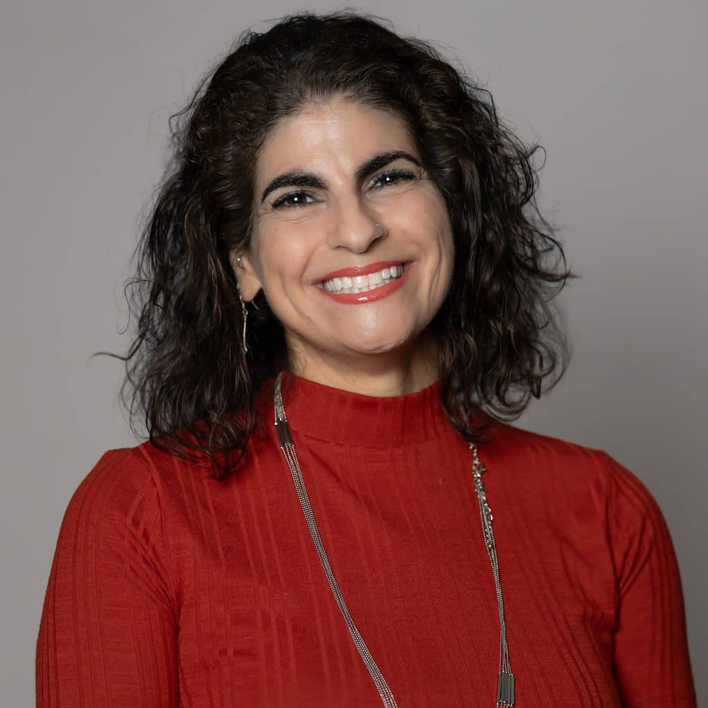 A smiling woman with dark, curly hair wearing a red mock-neck top and a long silver necklace.