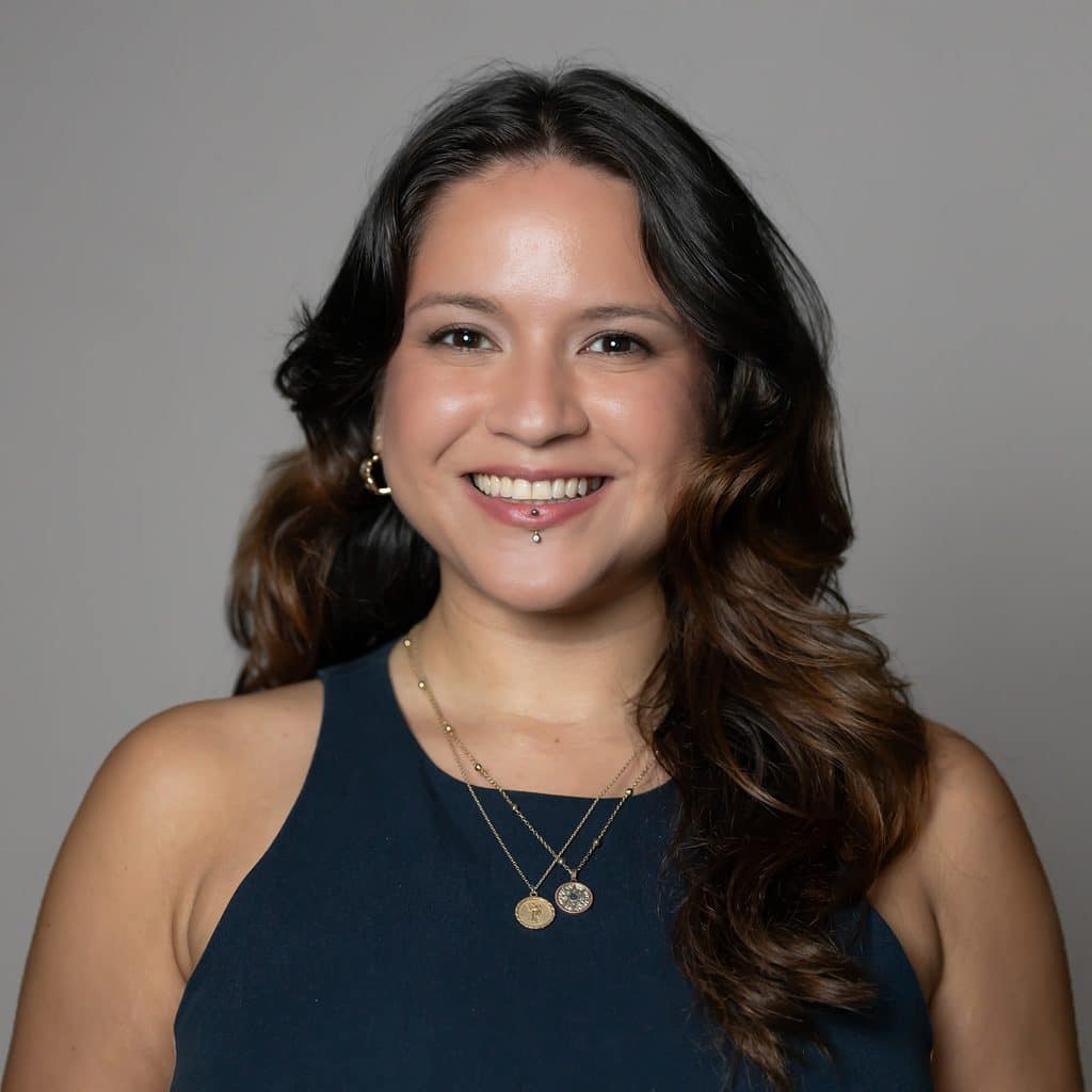 A smiling woman with a labret piercing wears a dark sleeveless top and two layered gold necklaces.