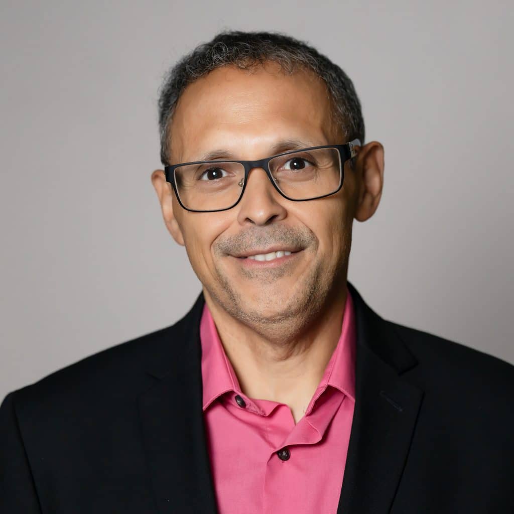 A man with short hair and glasses smiles while wearing a black blazer over a pink collared shirt.