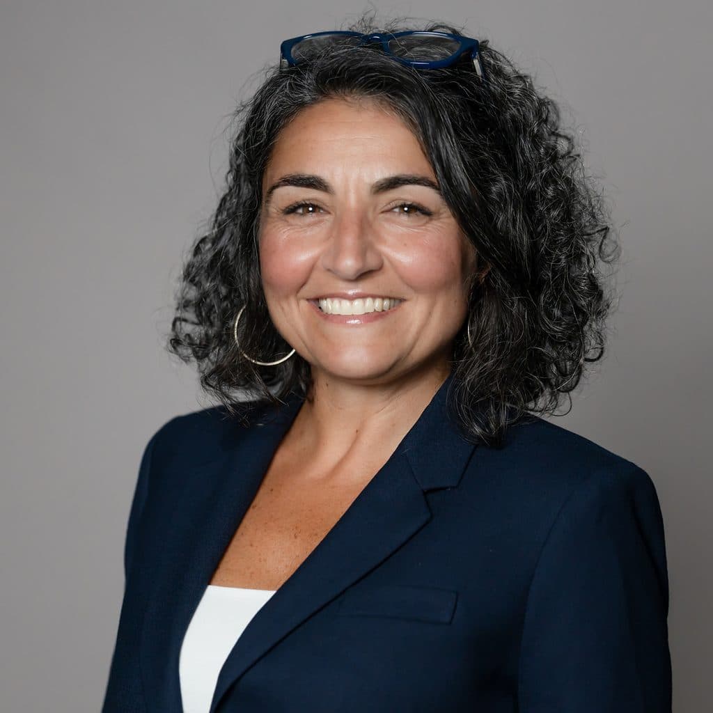 A woman with shoulder-length curly dark hair wearing a navy blazer and glasses atop her head smiles at the camera against a gray background.