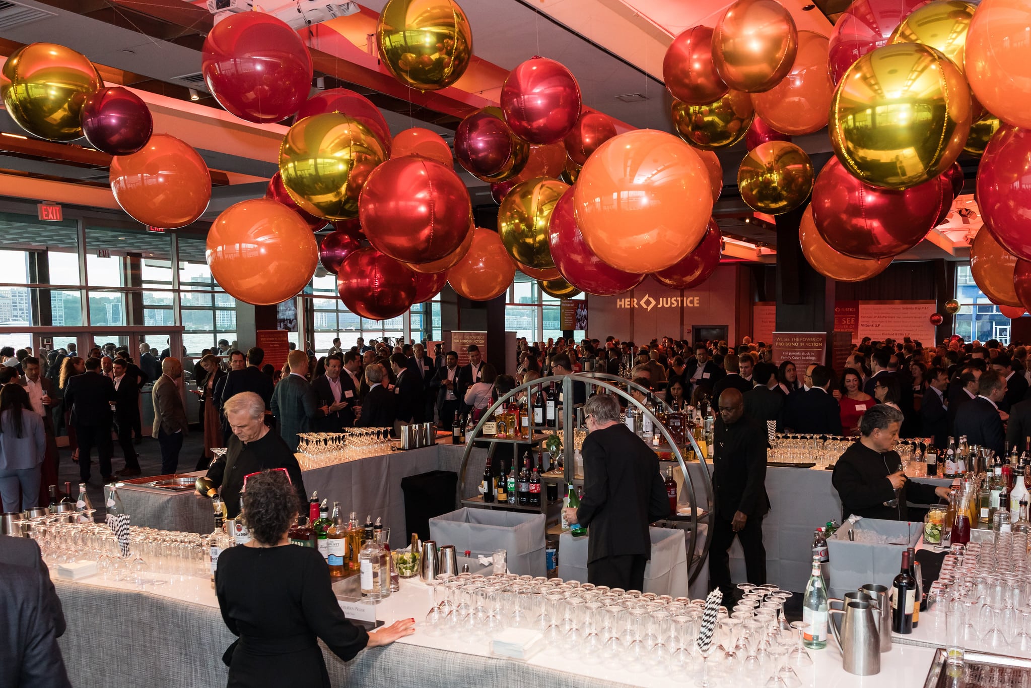 A large indoor event with many people mingling around a central bar area, decorated with large metallic balloons in red, orange, and gold. Bartenders prepare drinks, and glassware is arranged on the counters.