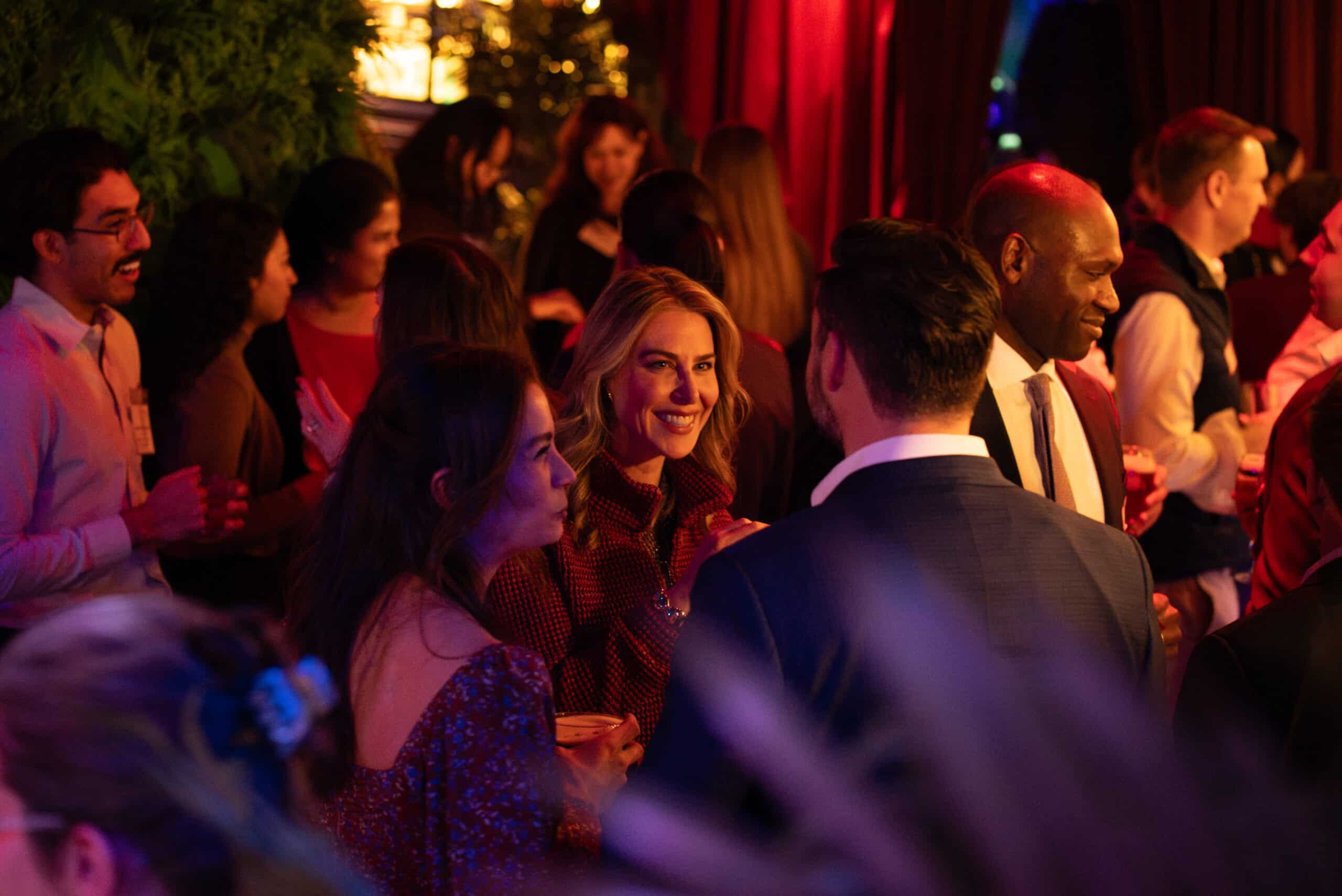 People are gathered at an indoor event. Most are dressed in semi-formal attire, talking and holding drinks. Warm lighting and red curtains create a festive atmosphere. One woman in the center is smiling at the camera.