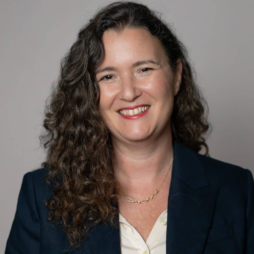 A smiling woman with long, curly dark hair wearing a navy blazer and a light-colored collared shirt.