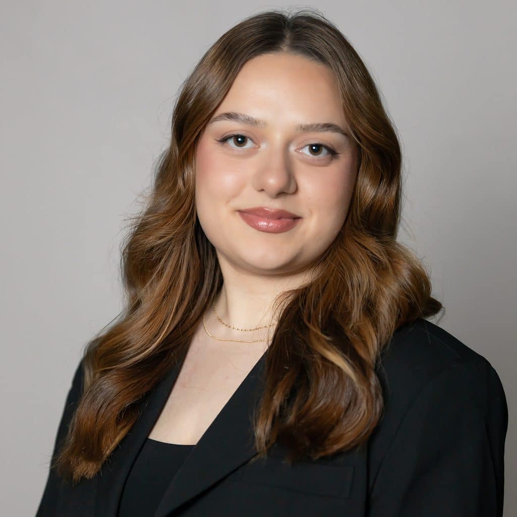 A woman with long, wavy brown hair wearing a black blazer looks at the camera.
